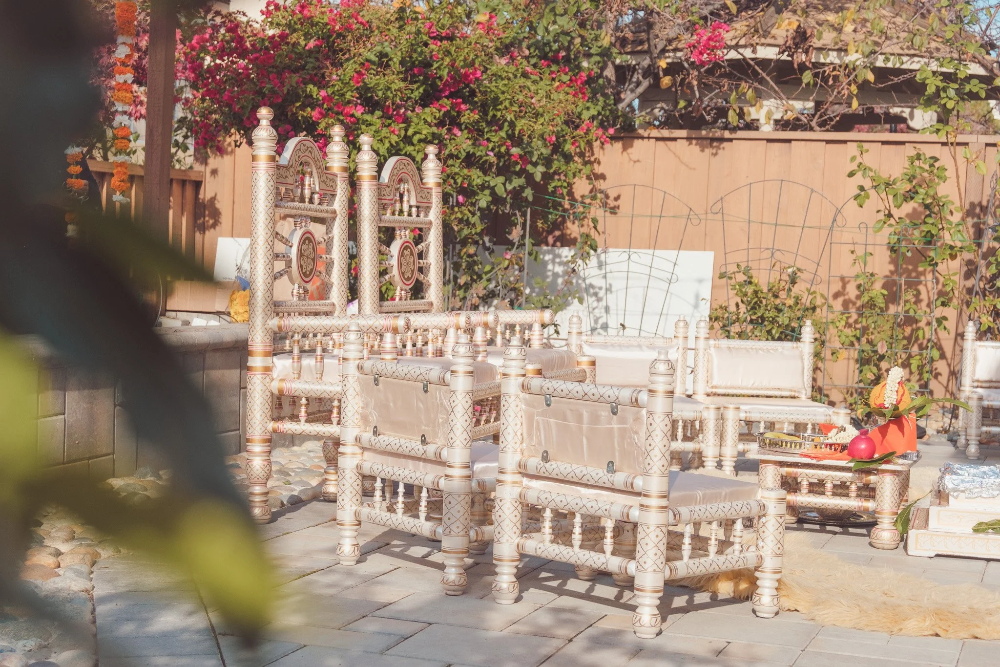 Outdoor patio with traditional Indian decorated chairs, pink flowers in the background, and a small decorated altar with flowers and offerings.