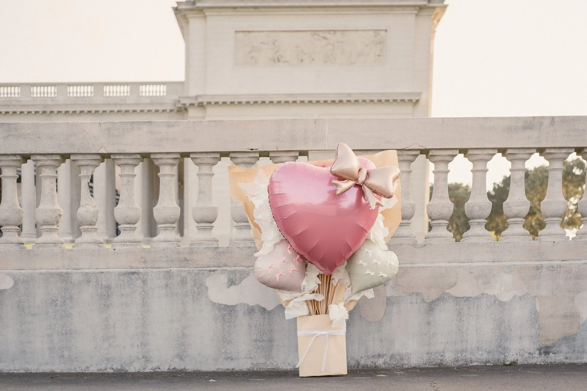 Pink heart-shaped balloon with a bow on top, surrounded by smaller balloons in soft pastel colors, against a stone railing and building background.