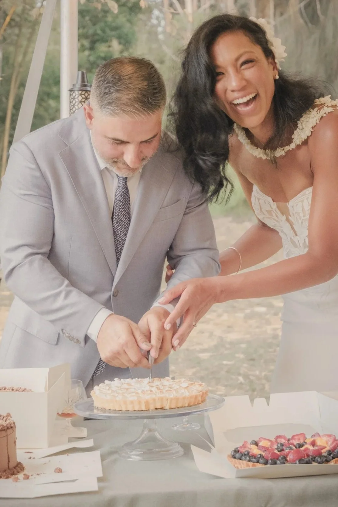 A woman and a man are cutting a wedding cake together and smiling, at an outdoor wedding reception.