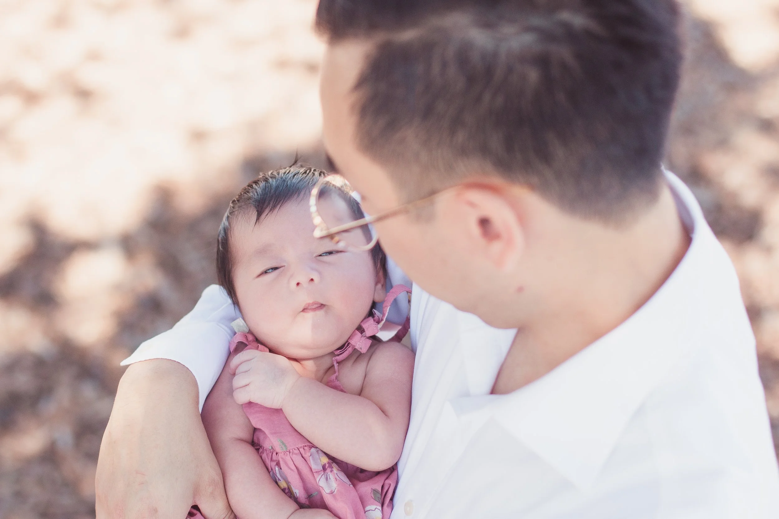 A man holding a baby girl outdoors, close-up view of their faces, with the man wearing glasses and a white shirt, and the baby in a pink dress, looking at each other.