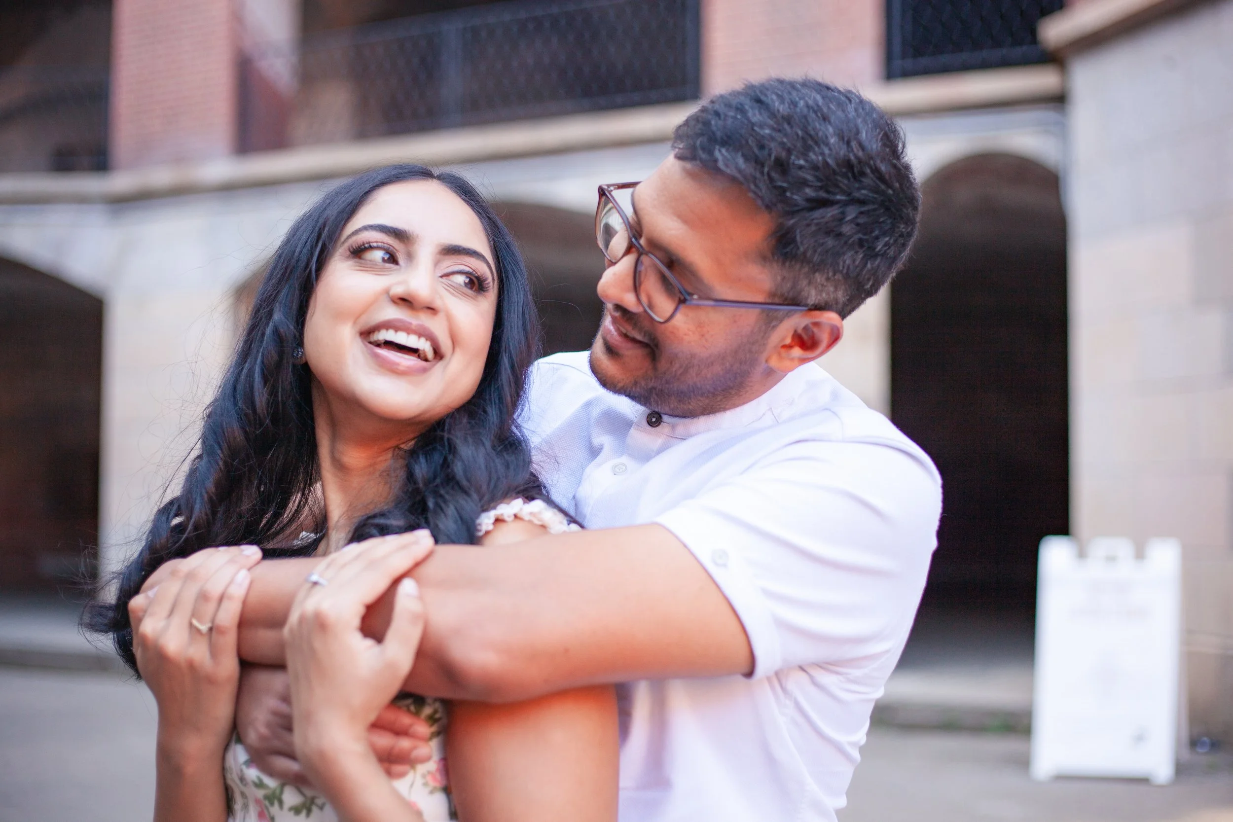 A man and woman smiling and embracing outdoors near a building.
