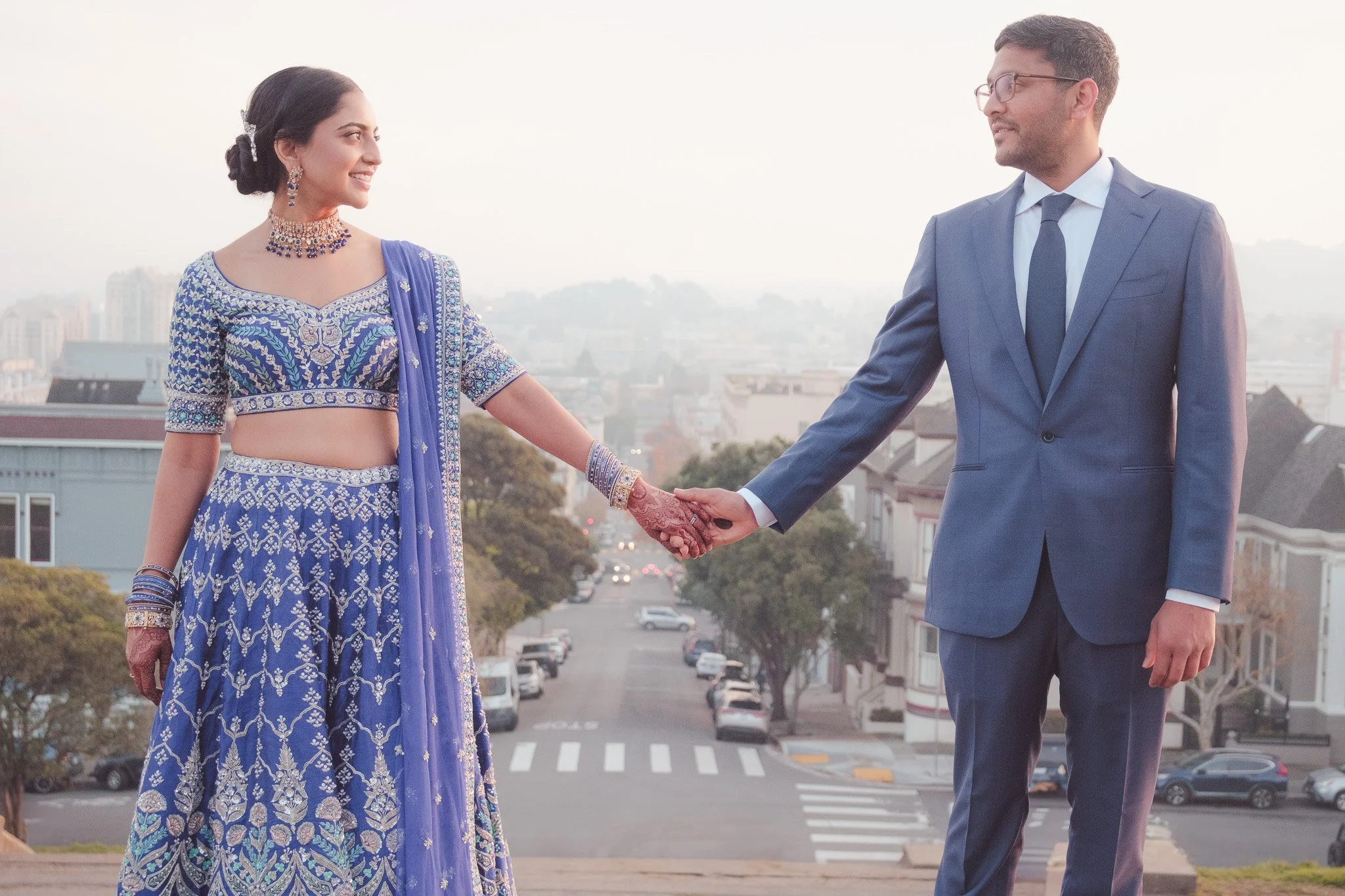 A woman in traditional Indian attire holding hands with a man in a blue suit on a city street during sunset.
