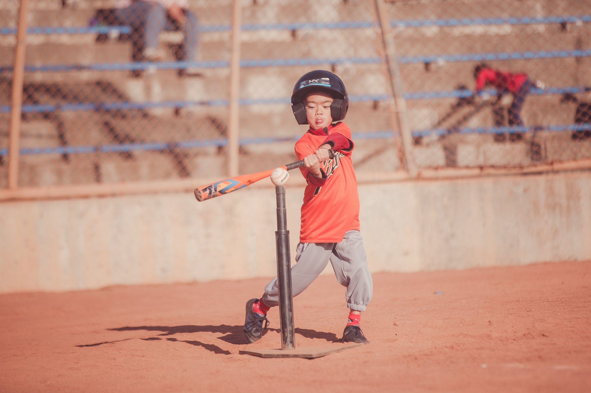 A young boy wearing a black helmet and red shirt practicing baseball with a bat on a clay field, with a chain-link fence and two other children in the background.