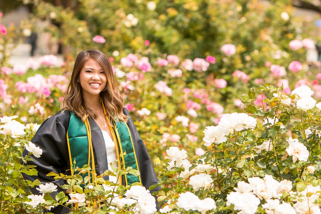 A smiling young woman in a graduation gown and cap standing in a garden of blooming roses.