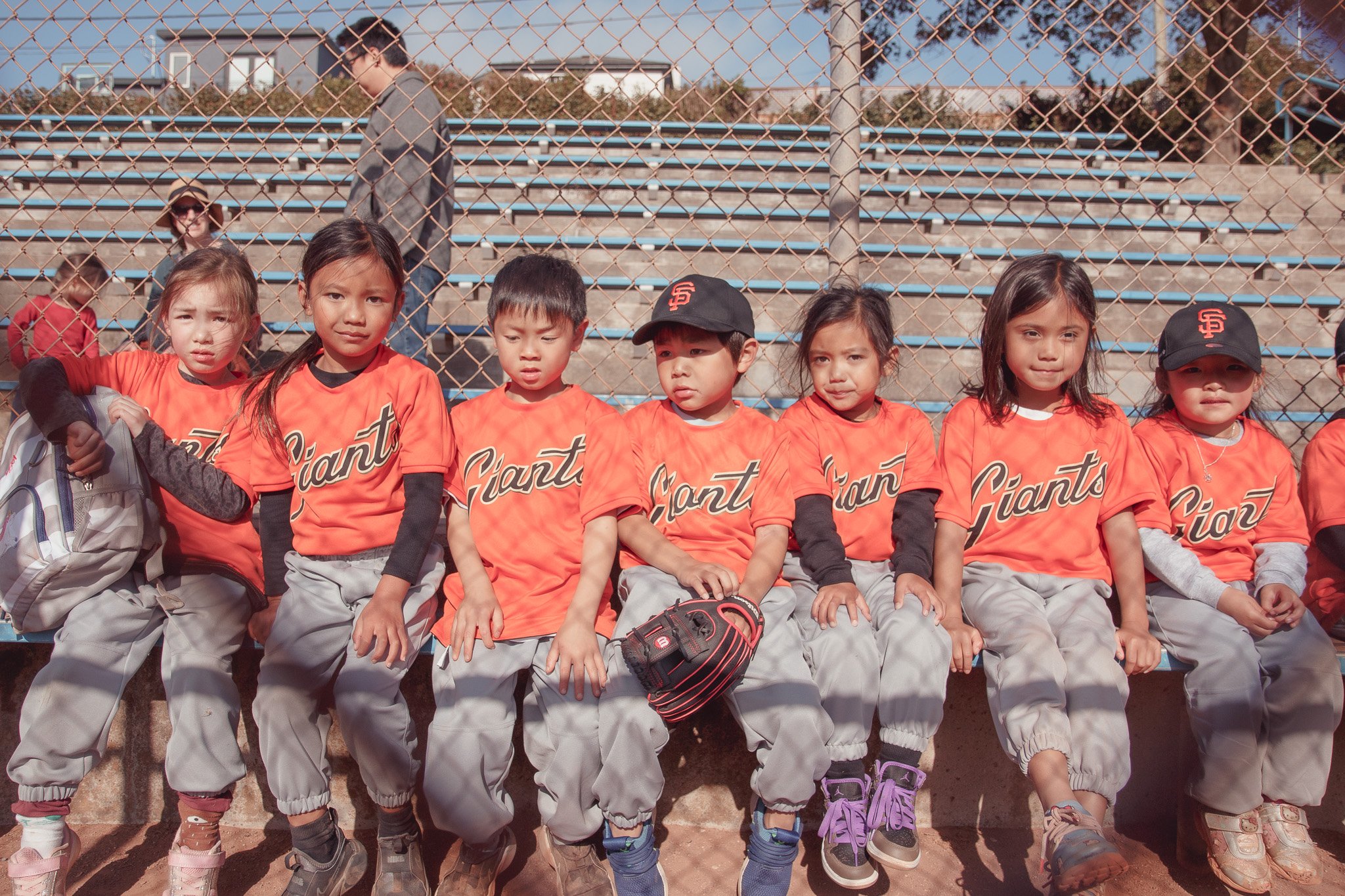 Youth baseball team sitting on a bench, wearing orange jerseys with 'Giant' written on them, with a chain-link fence and bleachers in the background.