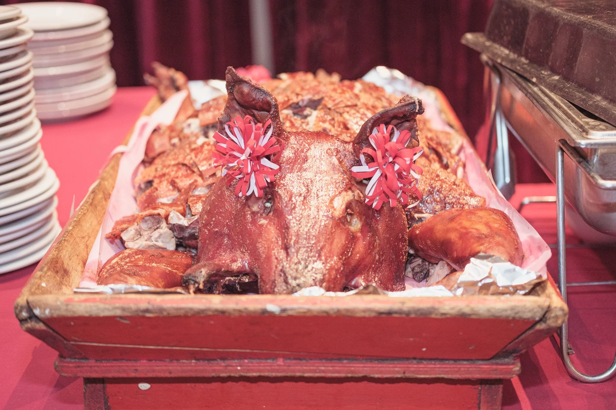 Cooked whole pig with decorative paper flowers on its ears, displayed on a wooden tray at a buffet