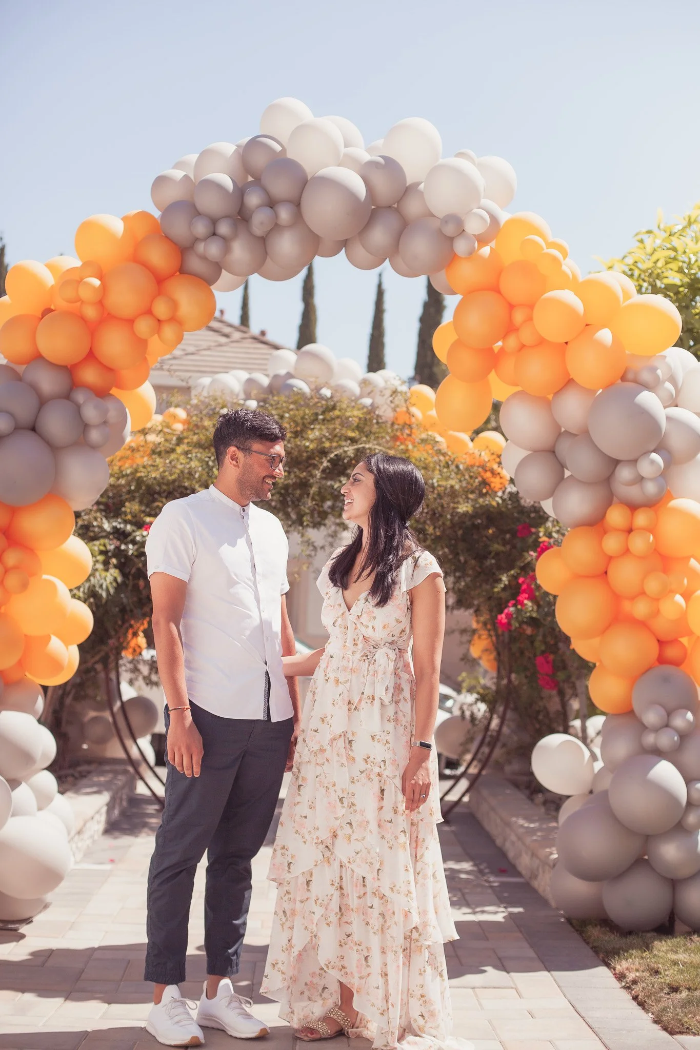A man and woman standing under a balloon arch at an outdoor event, smiling at each other.