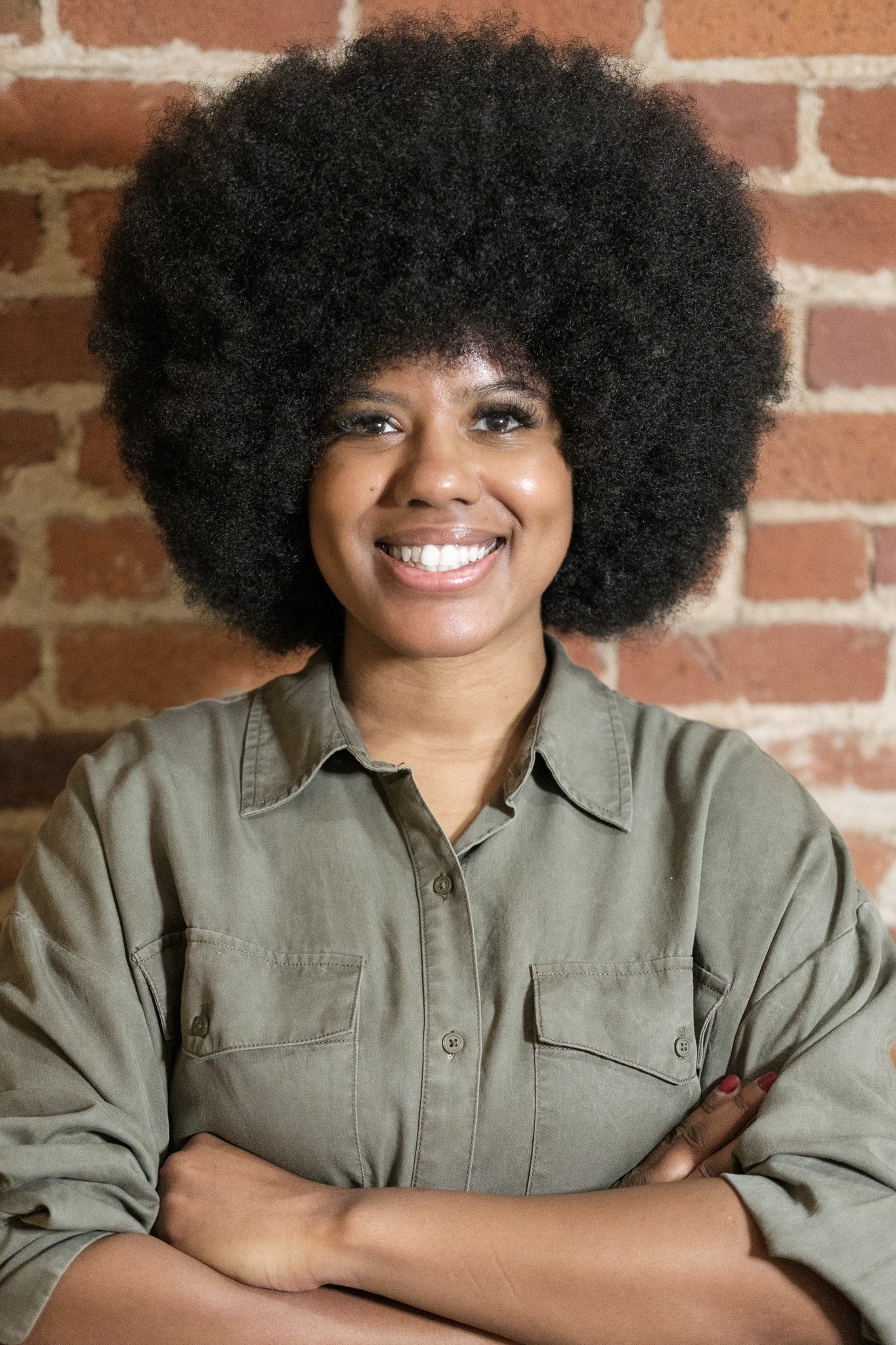 A woman with a large, textured afro hairstyle smiling with crossed arms in front of a brick wall.