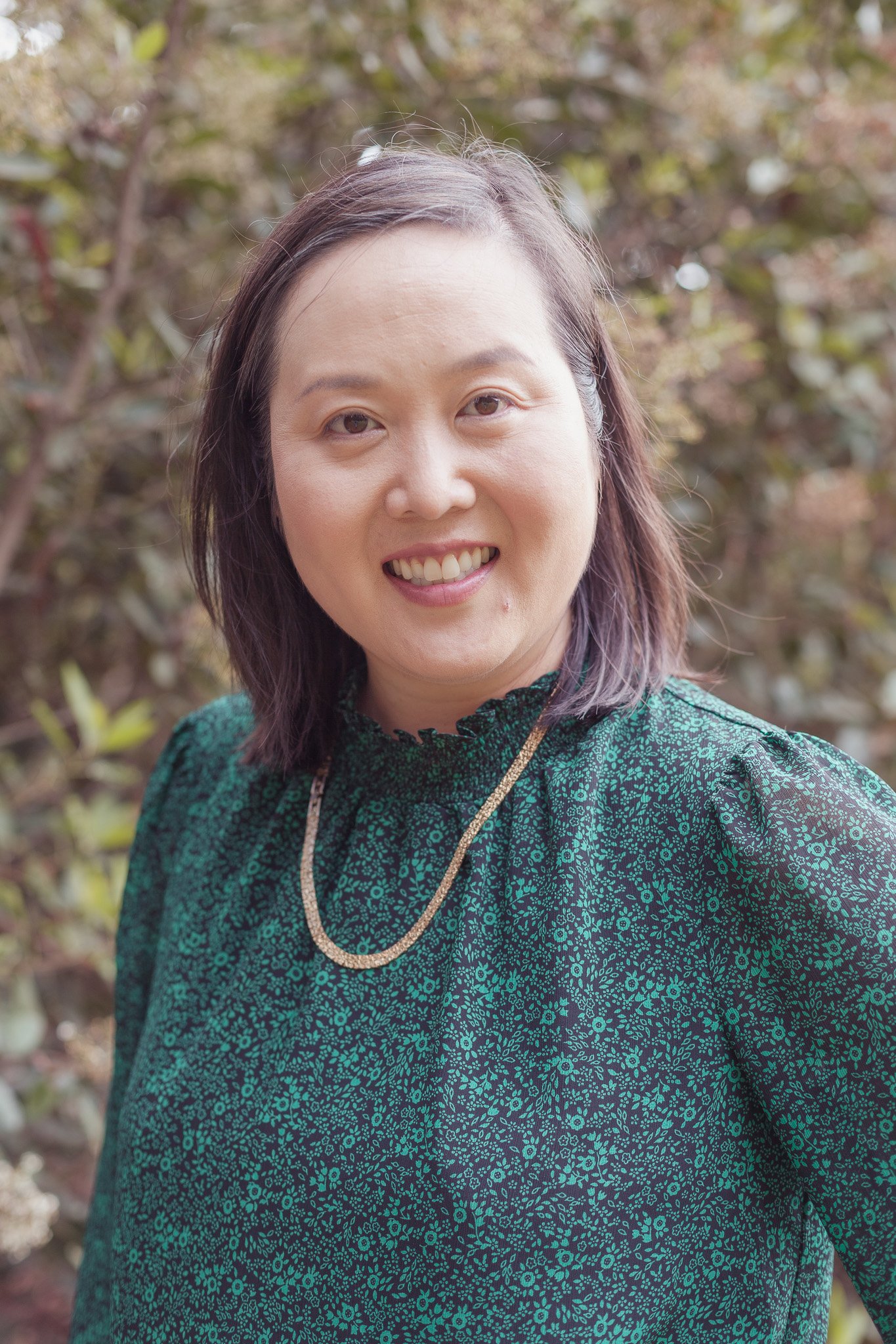 A woman with shoulder-length dark hair wearing a green patterned top and a gold necklace, smiling outdoors with trees in the background.