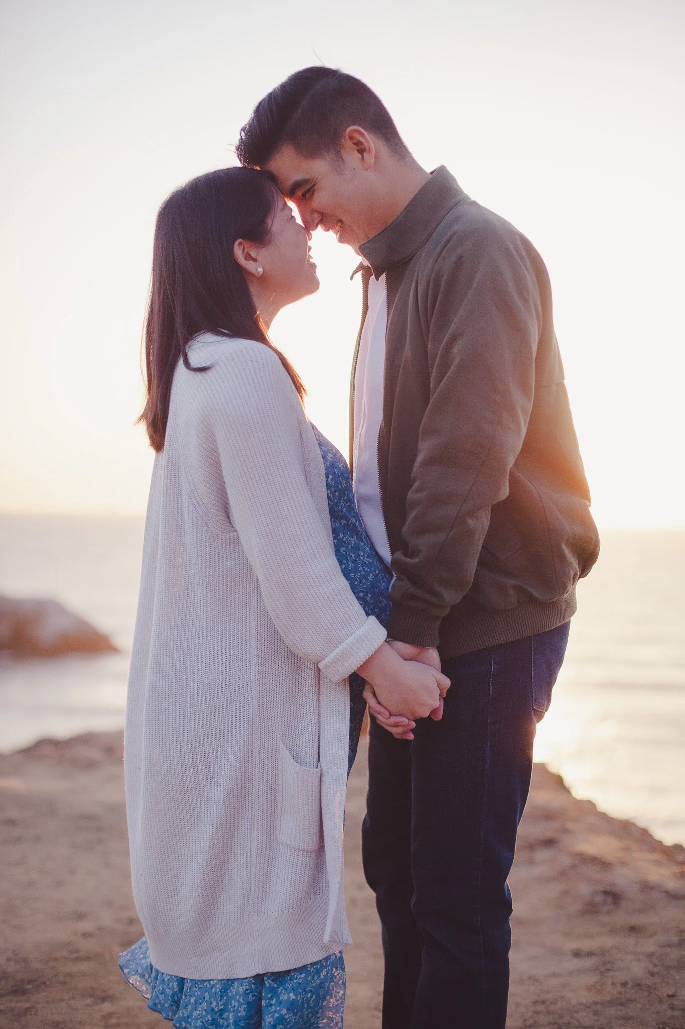A couple holding hands and smiling at each other on a beach during sunset.