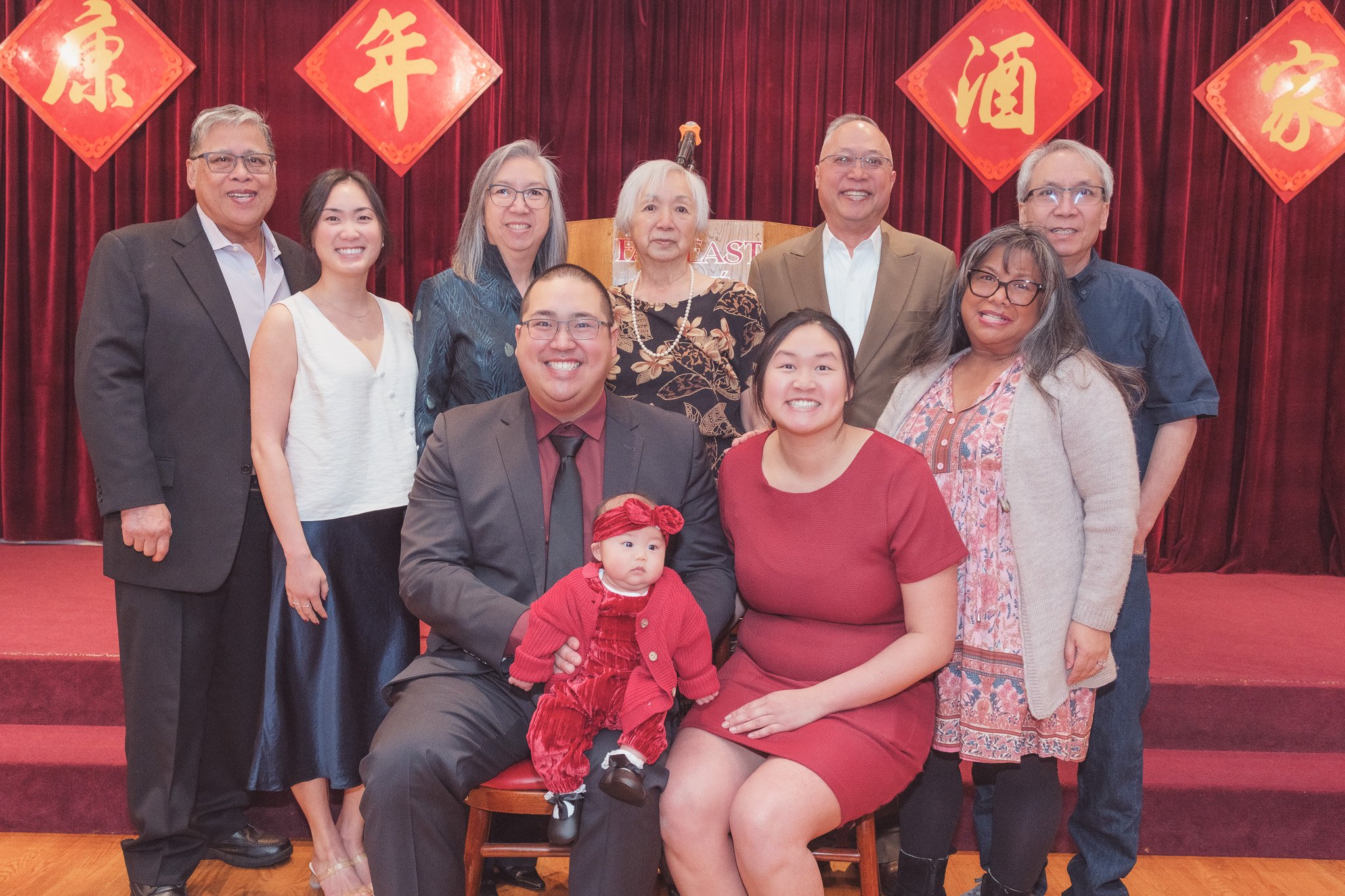 Group of eleven diverse people, including children and adults, posing on a stage with red curtains and Chinese New Year decorations behind them, including red diamond-shaped banners with Chinese characters.