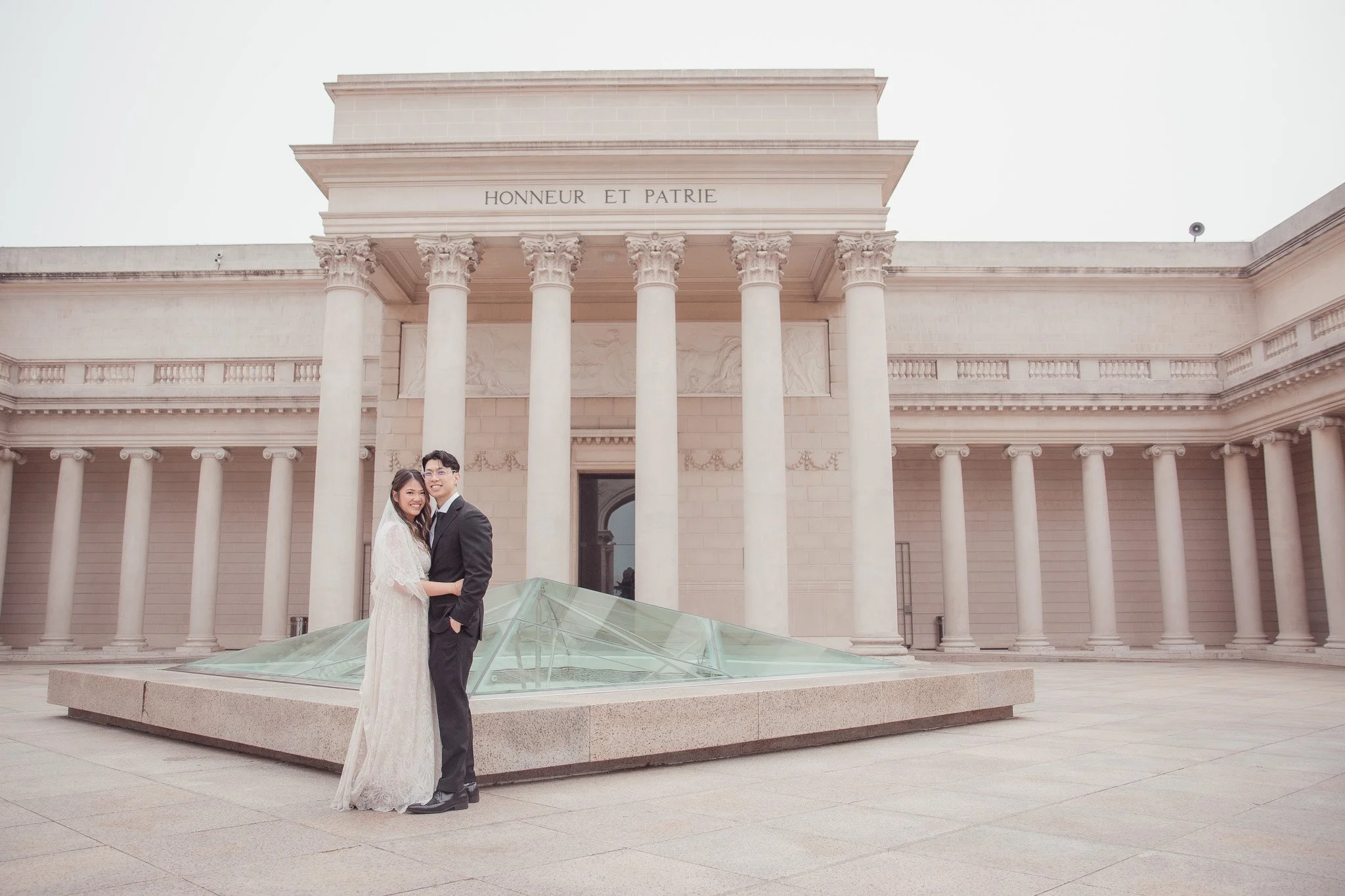 A newlywed couple in wedding attire stand embracing in front of a neoclassical building with tall columns and the inscription "HONNEUR ET PATRIE"