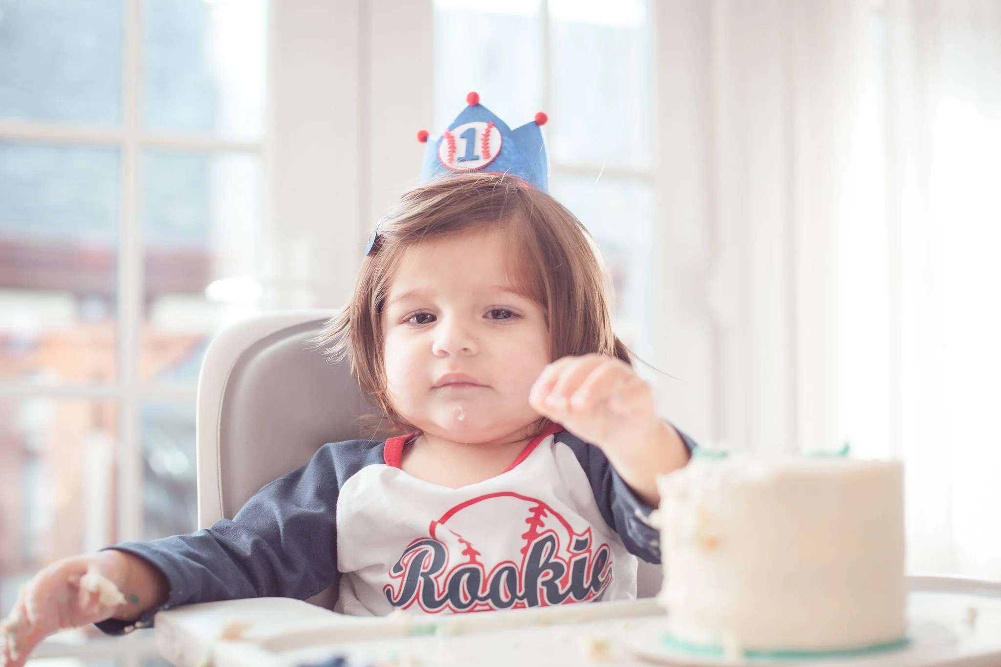 Young girl with brown hair sitting in a high chair, wearing a birthday hat with the number 10, and a shirt that says "Rookie". She is celebrating her birthday with a cake in front of her.