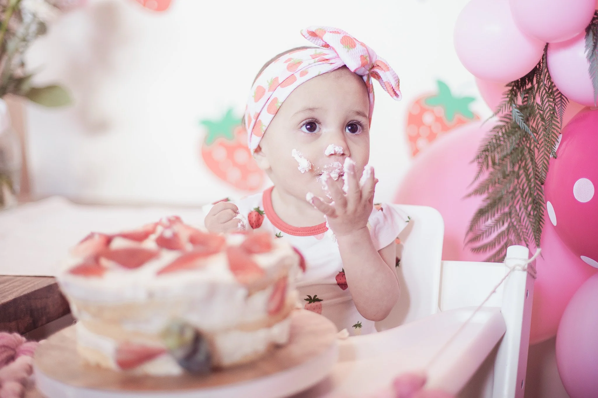 A young girl at her birthday party, wearing a strawberry-printed headband and shirt, with a small piece of cake on her face, sitting in a high chair surrounded by pink balloons and strawberry-themed decorations.