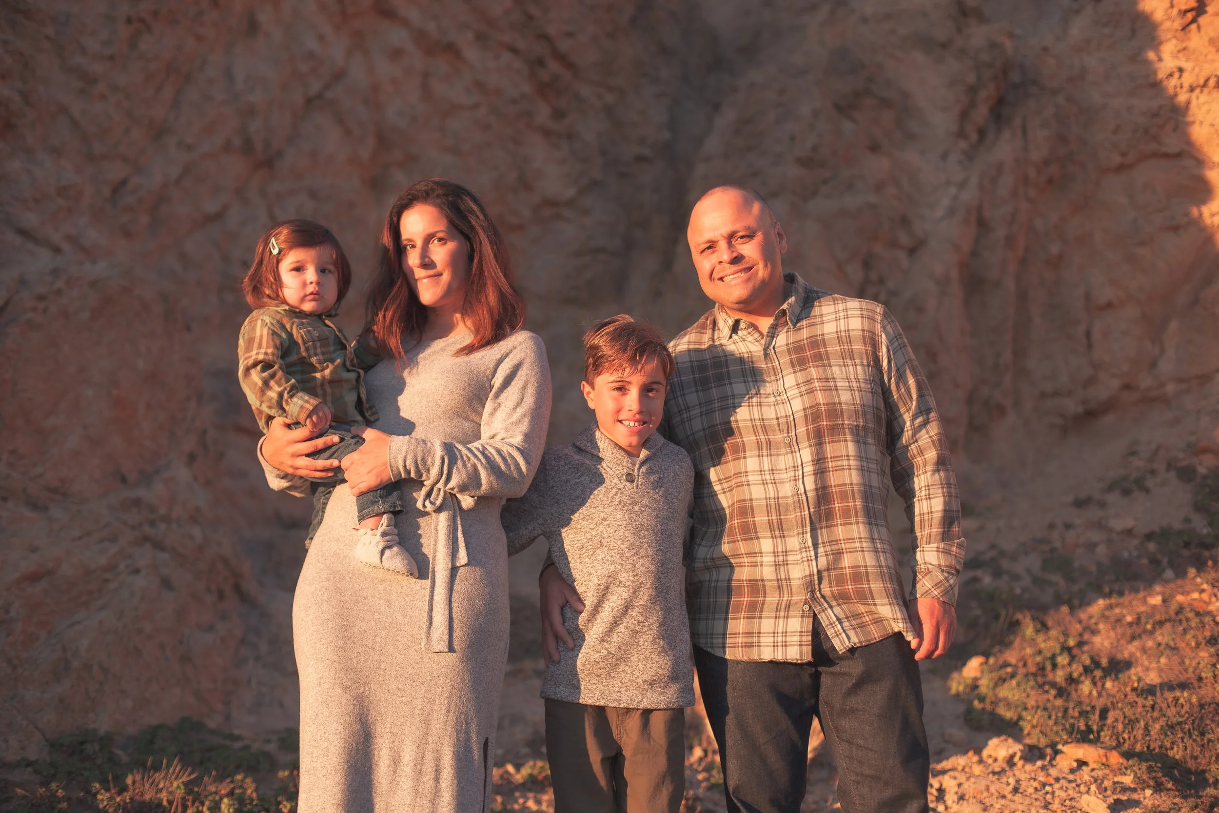 A family of five, including a woman, a man, and three children, standing outdoors with a rocky hill in the background during sunset.