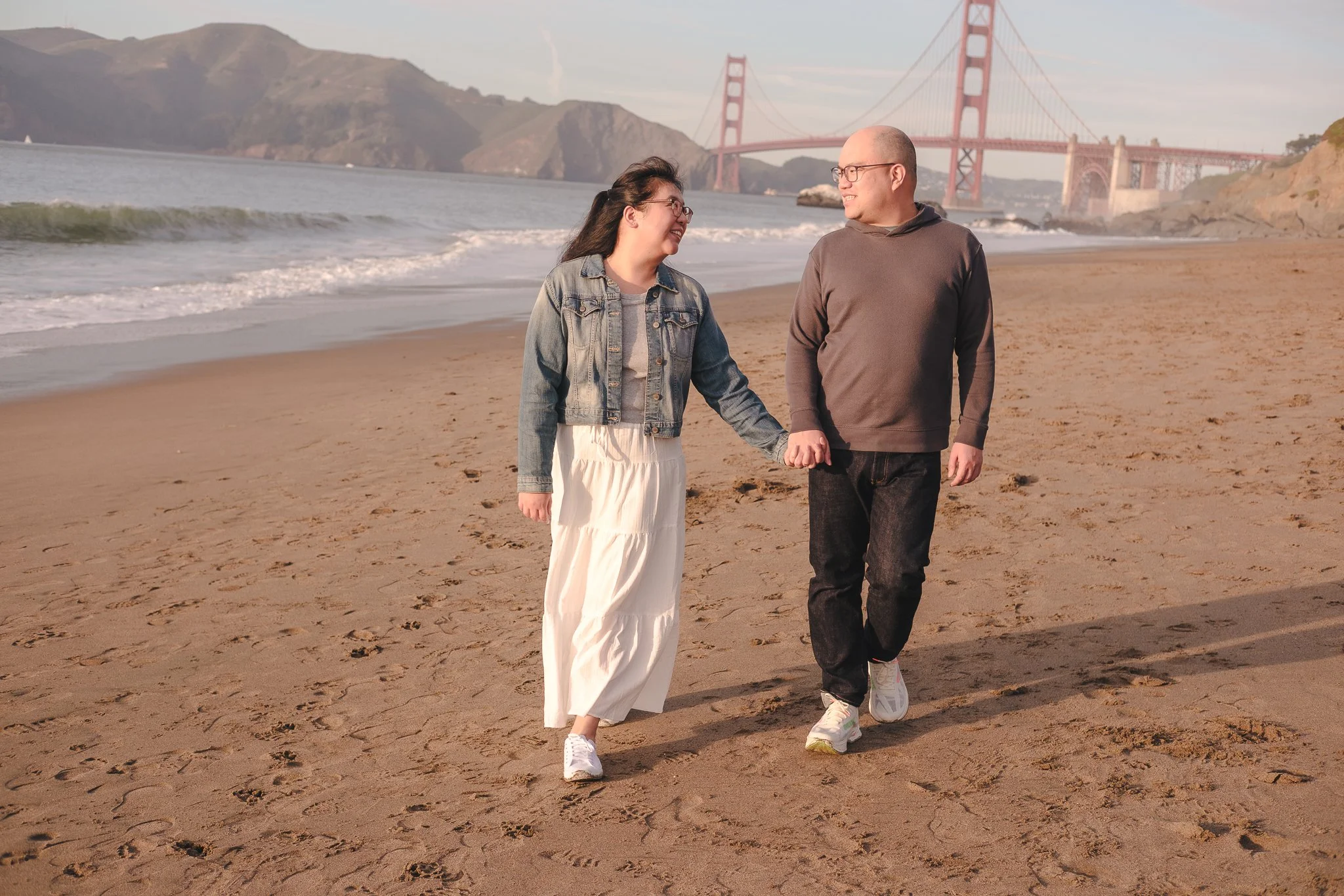 A couple holding hands and walking on a sandy beach during sunset with the Golden Gate Bridge in the background.