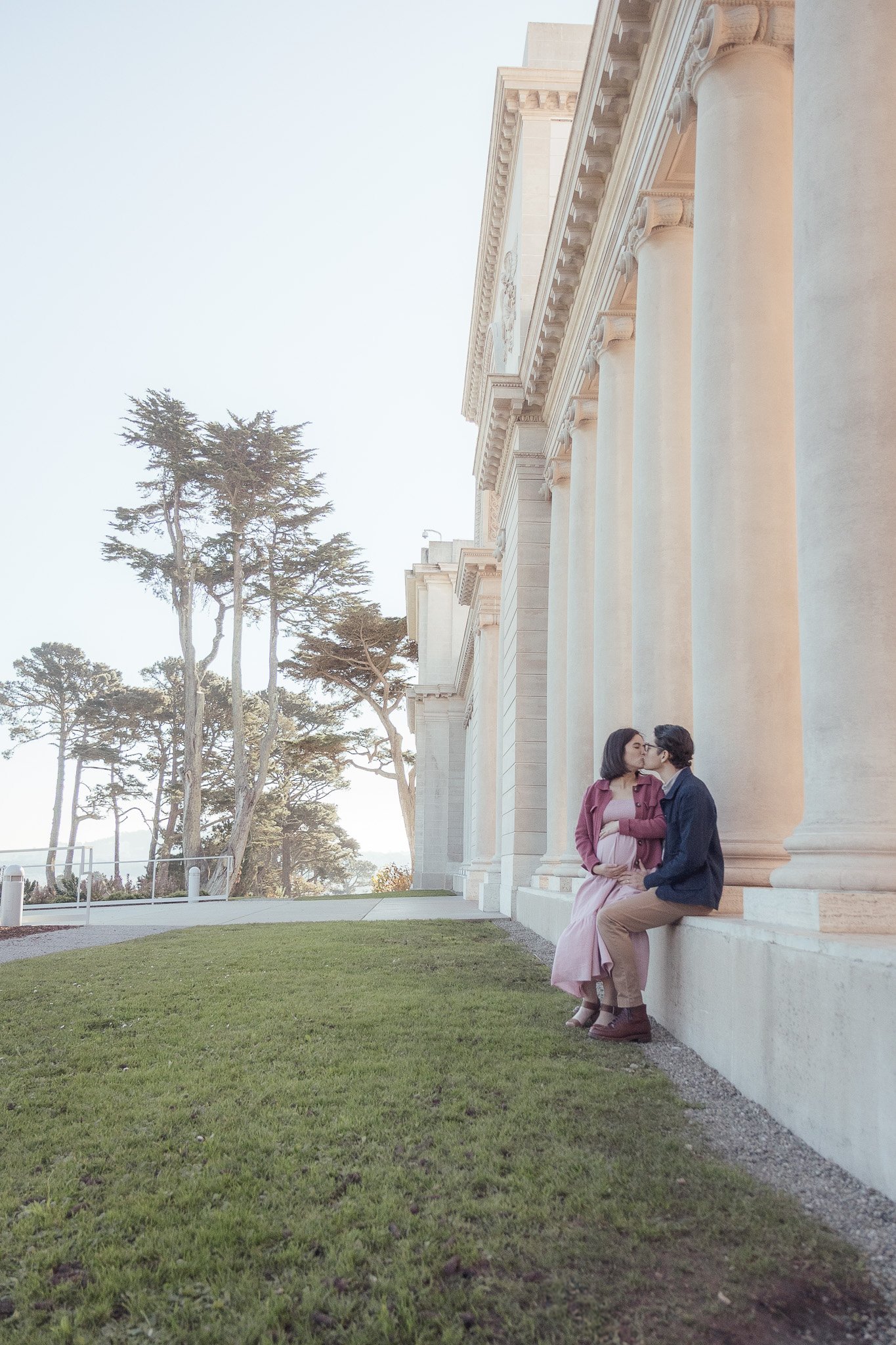 A couple sitting on a ledge kissing outside a historic building with large columns and trees in the background.