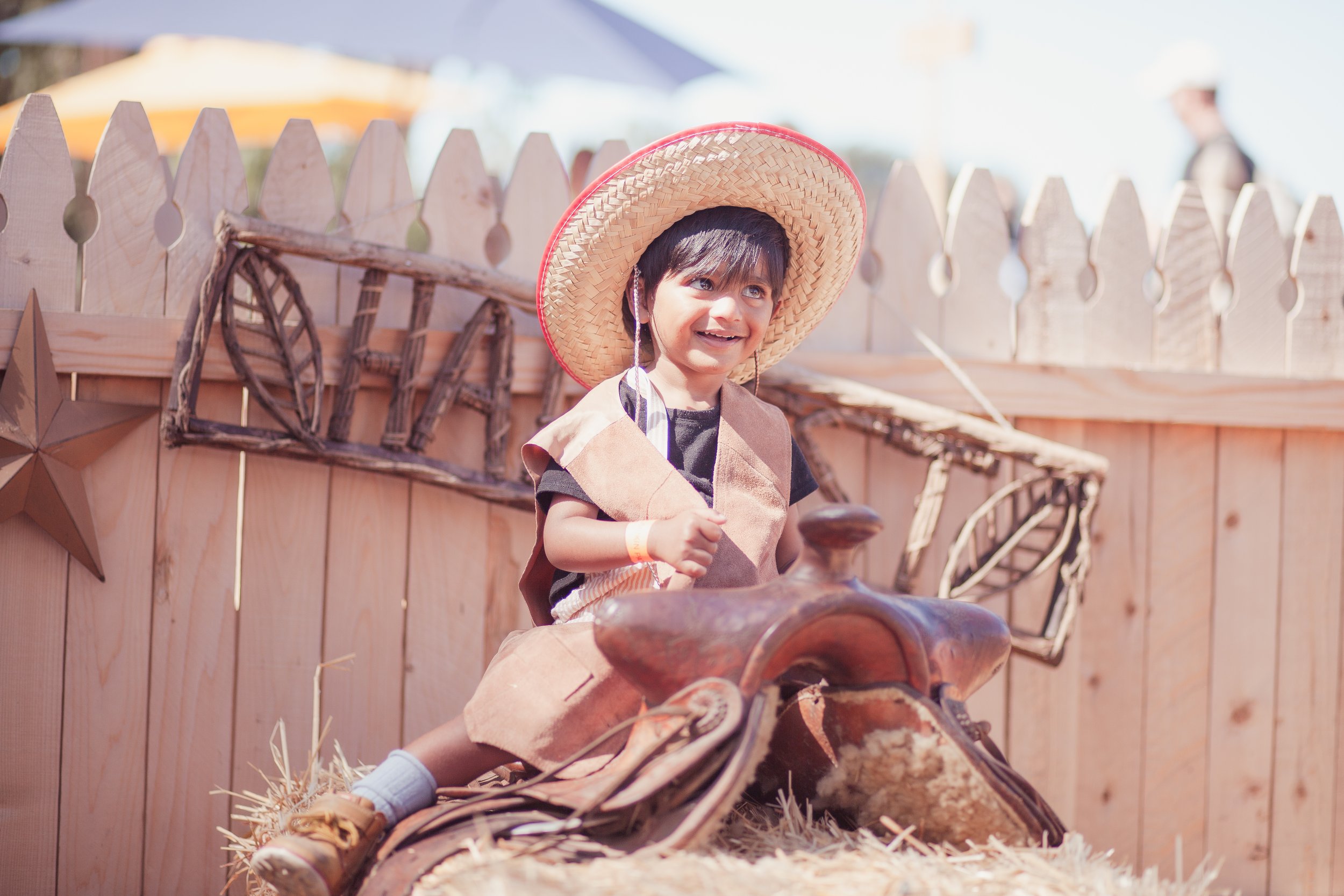 Young boy wearing a large straw hat and cowboy vest sitting on a saddle in a farm-like setting with wooden fence and decorative star in the background.