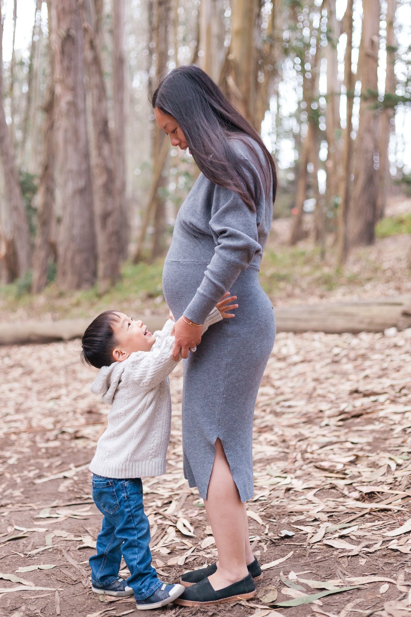 A pregnant woman bending down in a forest, smiling, while a young boy holding her hands looks up at her and smiles.