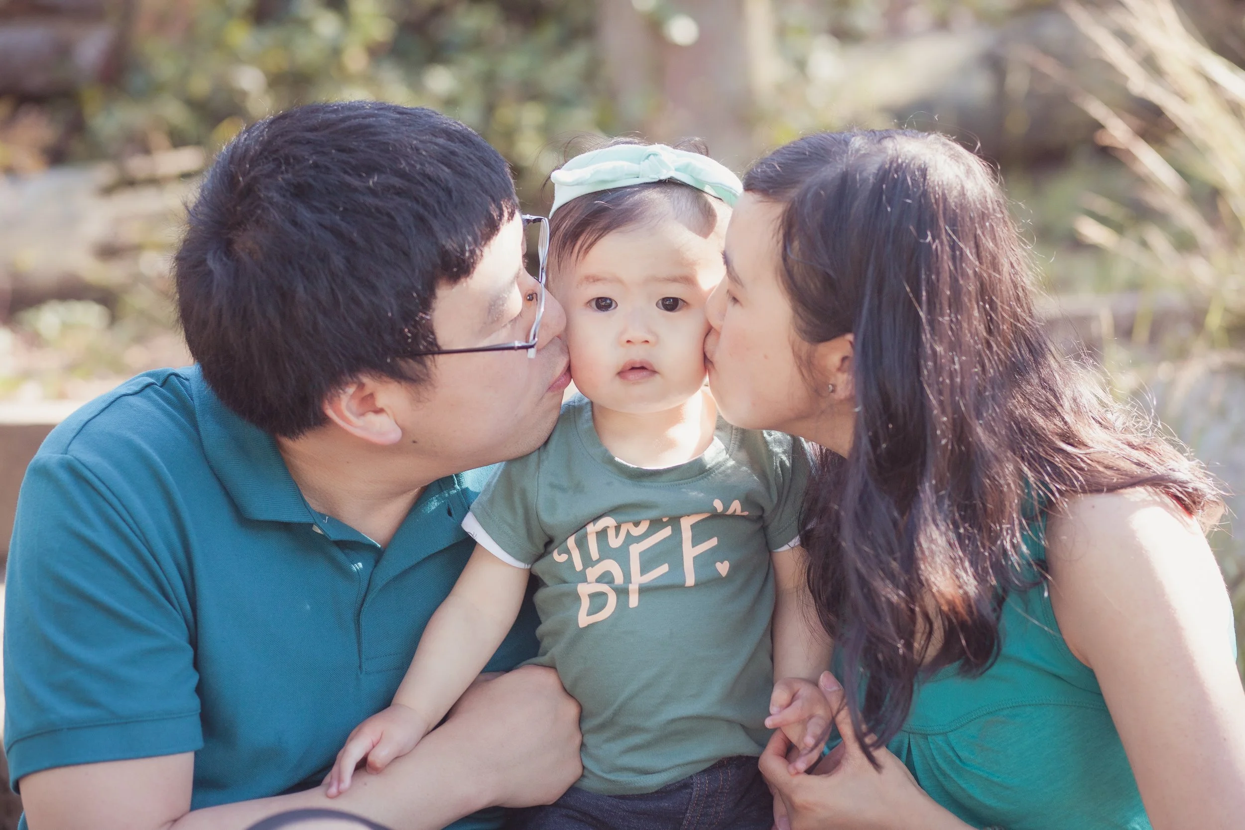 A young child being kissed on each cheek by a man and a woman outdoors, with trees in the background.