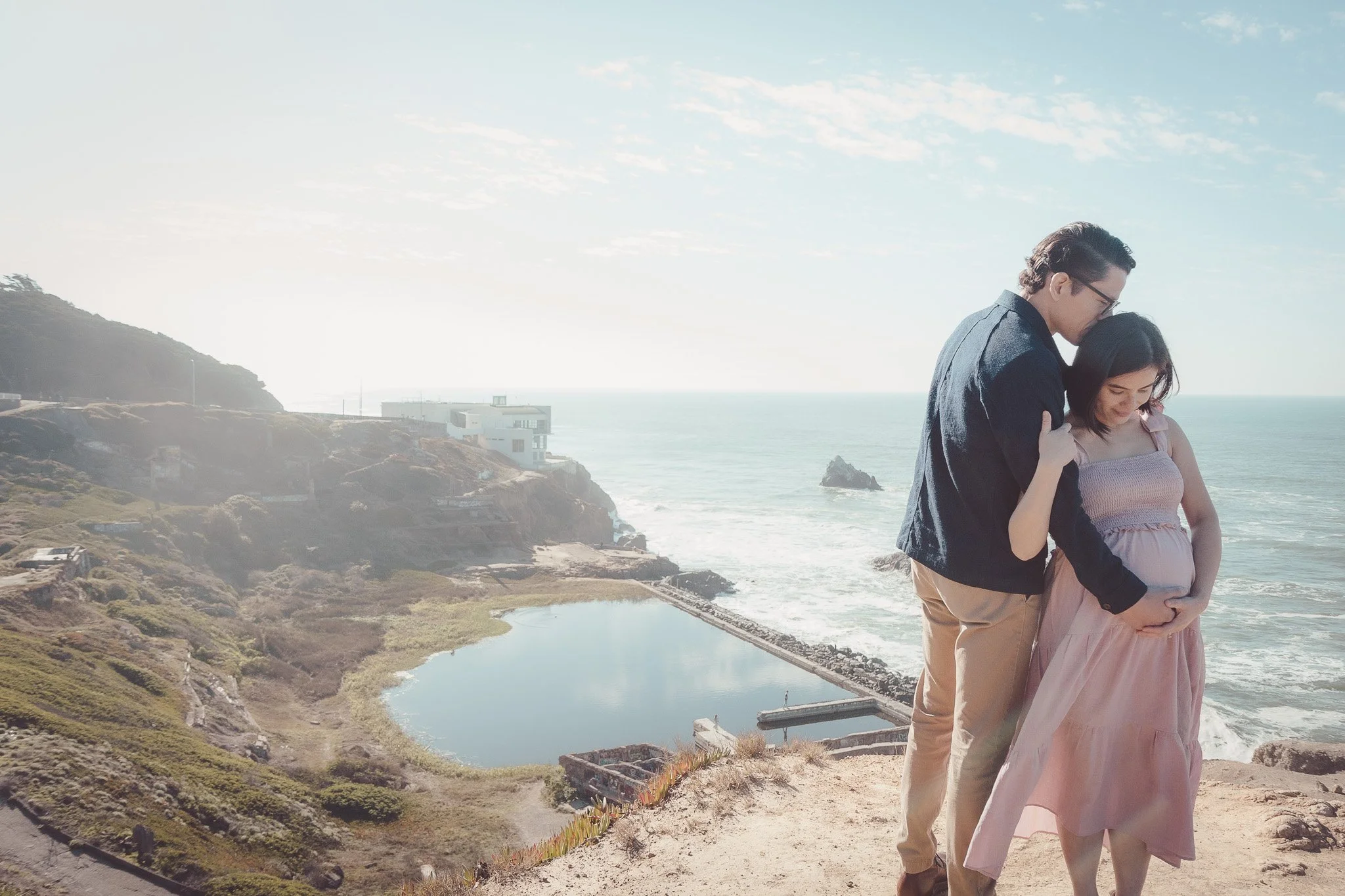 A couple, with the man holding the pregnant woman, standing on a beach overlooking the ocean and a pond, with houses on a hill in the background.