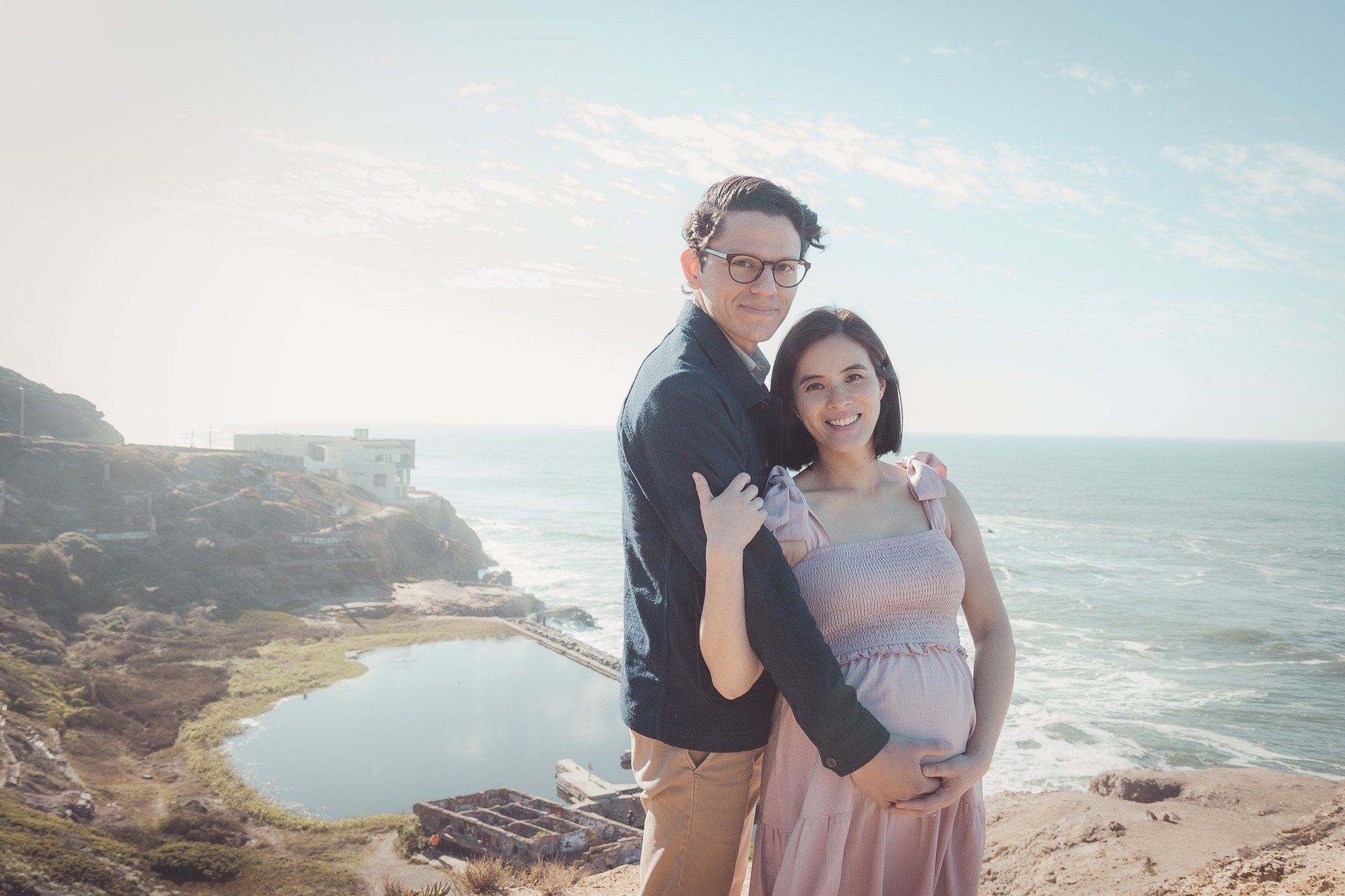 A couple standing on a cliffside with the ocean in the background, the pregnant woman is holding her belly, and the man has his arm around her, both smiling.