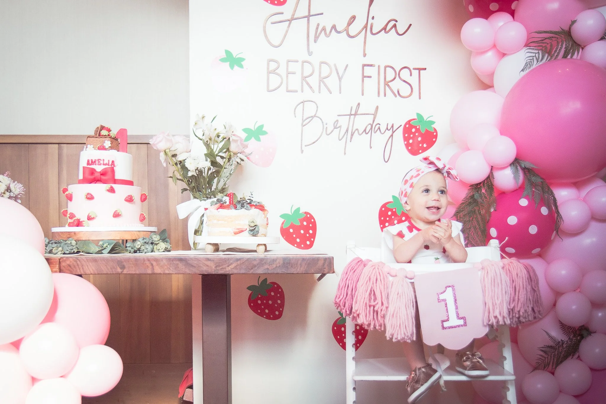 A young girl celebrating her first birthday, seated in a high chair decorated with a pink fringe and a number '1' banner, surrounded by pink and white balloons, a strawberry theme, a birthday cake with strawberries, and a wall sign reading 'Amelia Be