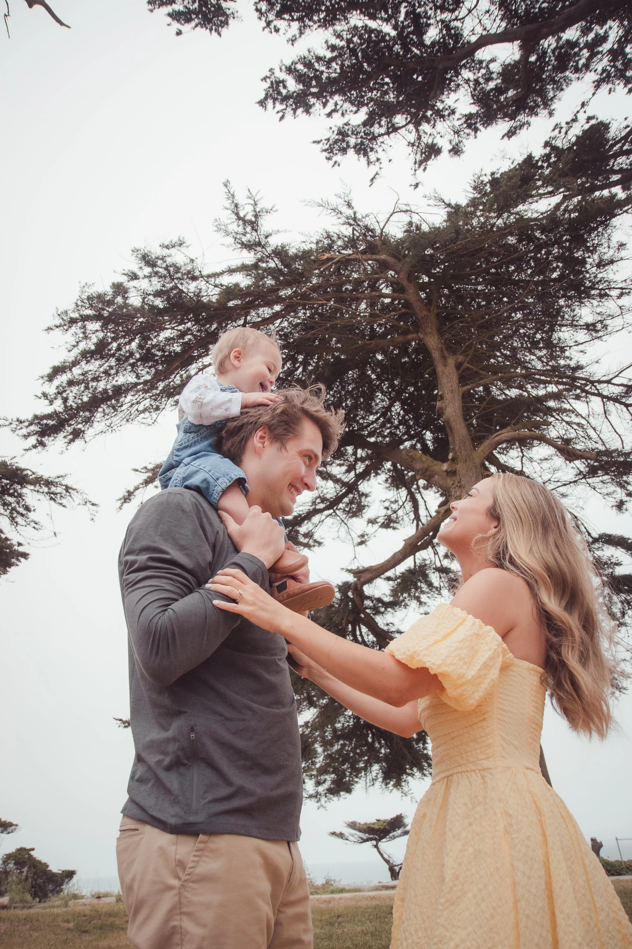A smiling woman in a yellow dress and a man in a gray shirt with beige pants hold a young child on his shoulders outdoors near a large tree, with a cloudy sky in the background.