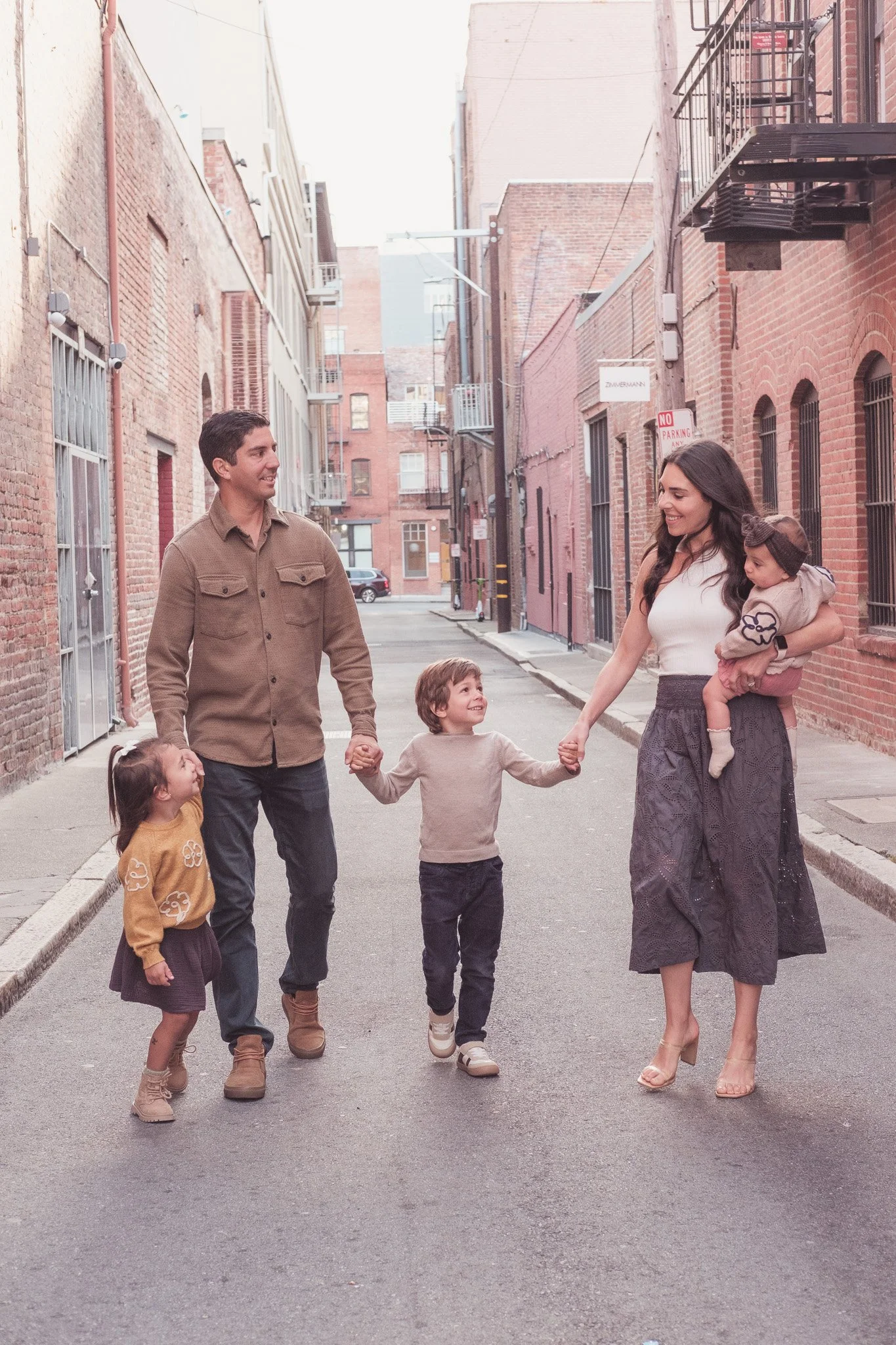 A family of four walking hand in hand on a city street, smiling and enjoying each other's company. The father on the left, the mother on the right, with two children between them. The street is lined with brick buildings and fire escapes.