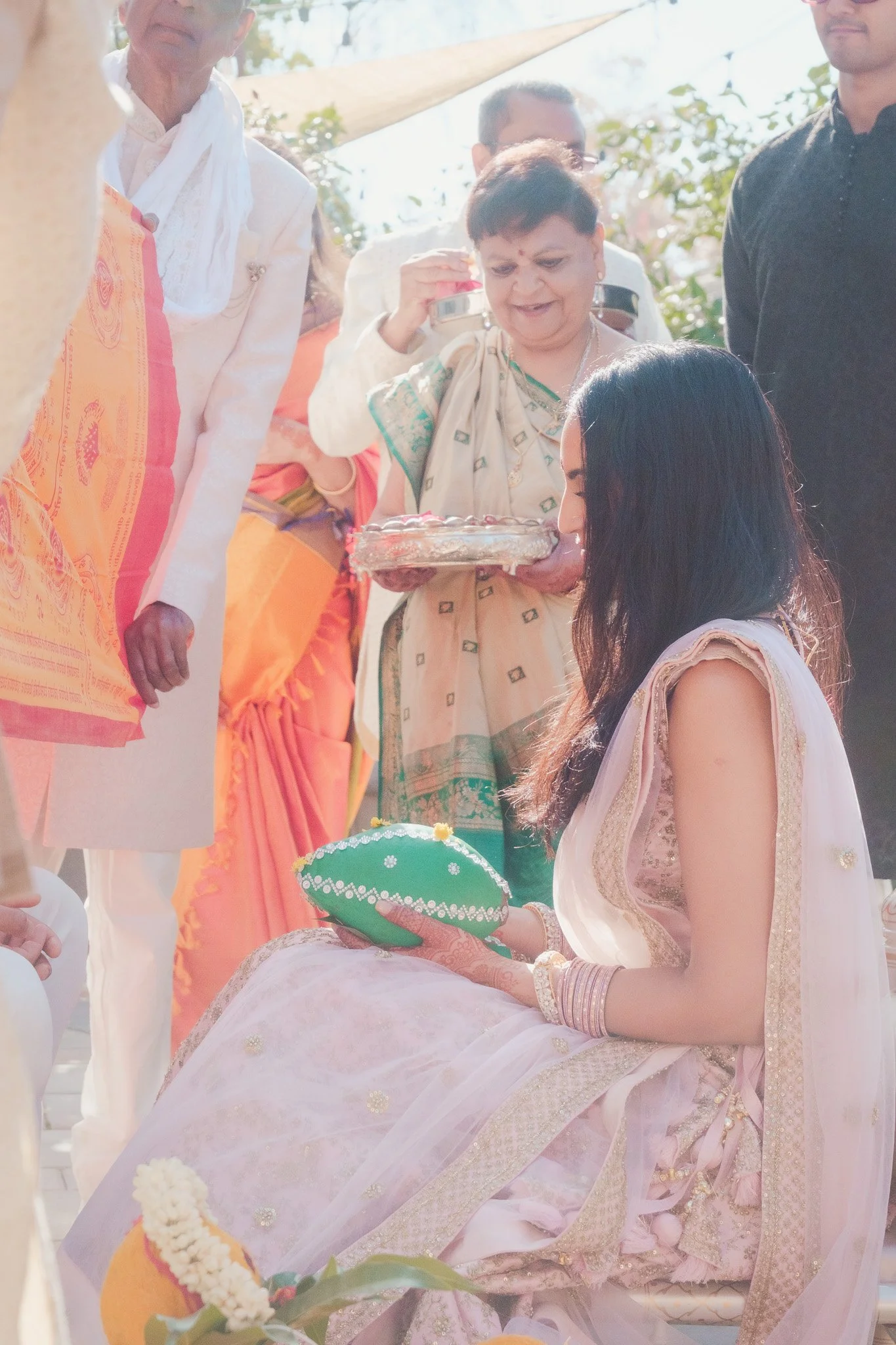 A woman in traditional Indian attire, sitting outdoors, holding a decorated green cushion, participating in a cultural ceremony with several other people around her.