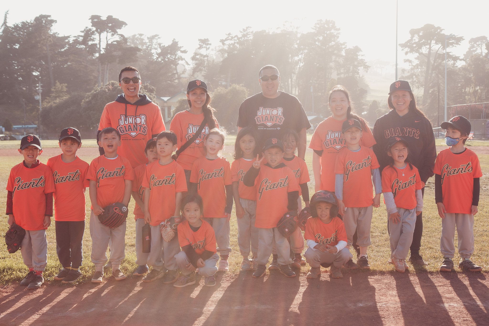A group of young children and coaches in orange and black San Francisco Giants baseball uniforms posing on a baseball field in bright sunlight.