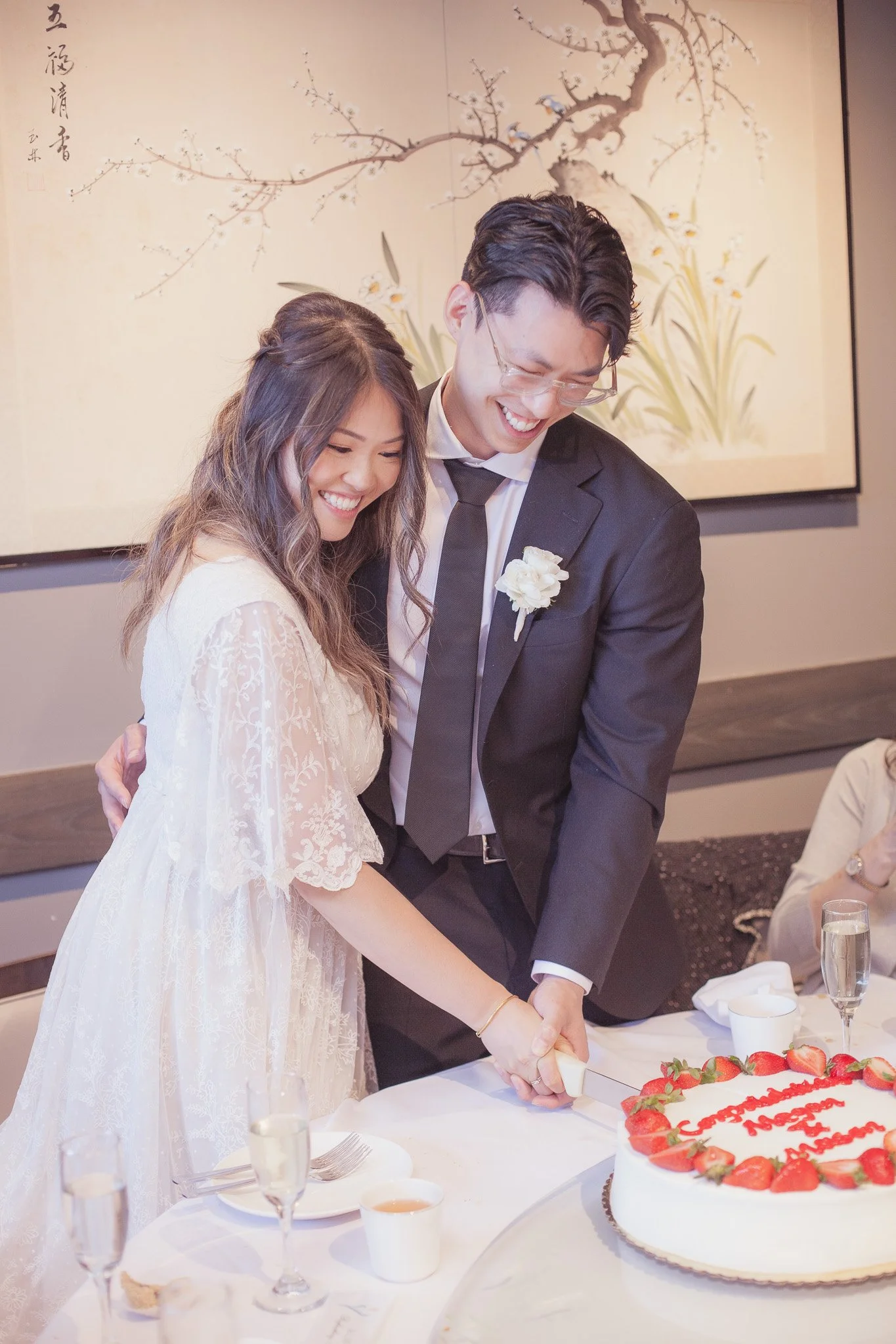 A couple is cutting a wedding cake together, smiling happily. The woman is wearing a white lace dress, and the man is dressed in a black suit with a white shirt and tie, with a white flower on his lapel. The cake is decorated with strawberries and ha
