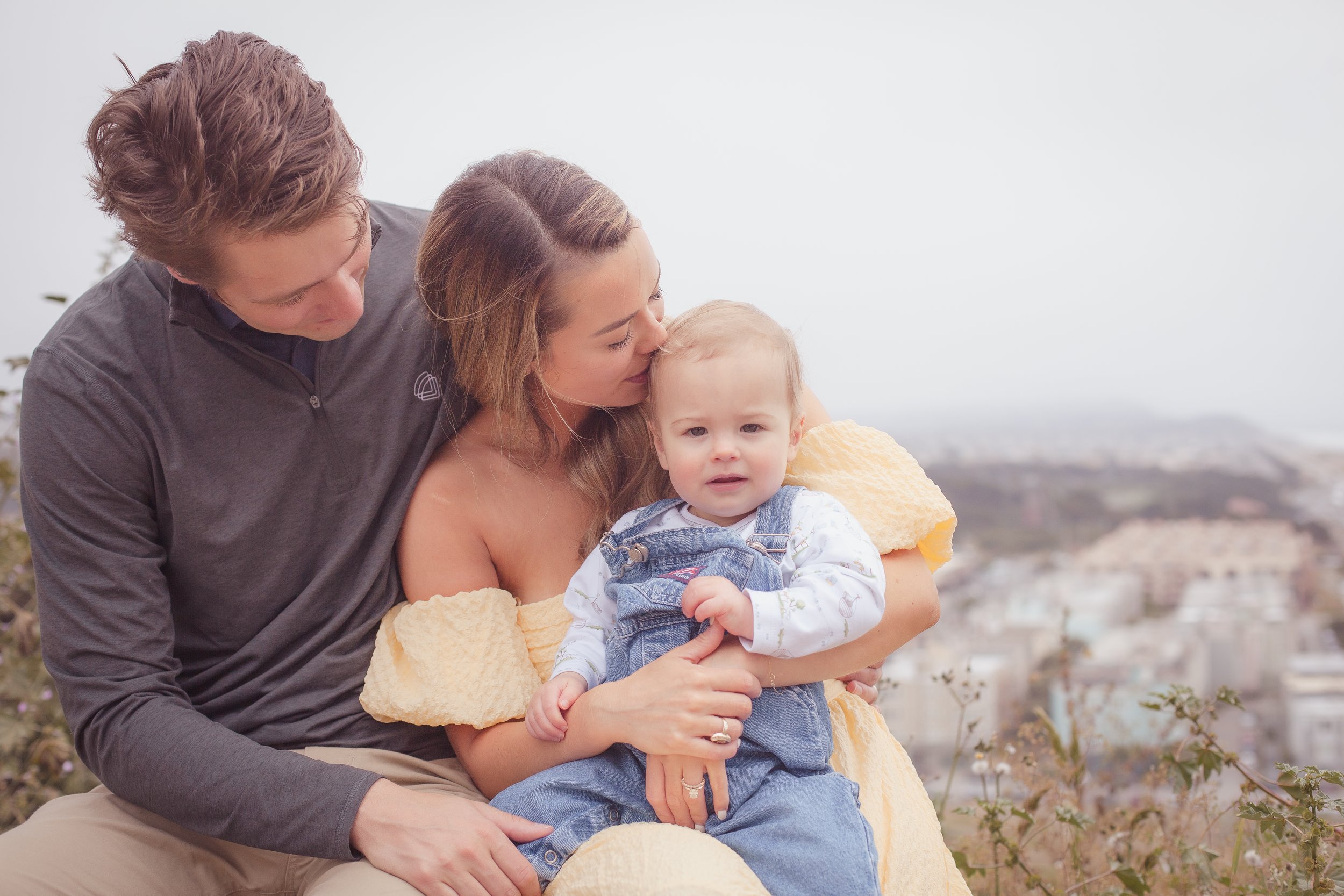 A family of three on a hilltop with a city view in the background. The mother is holding a young boy on her lap, and the father is sitting close, looking at the child, with a cloudy sky overhead.