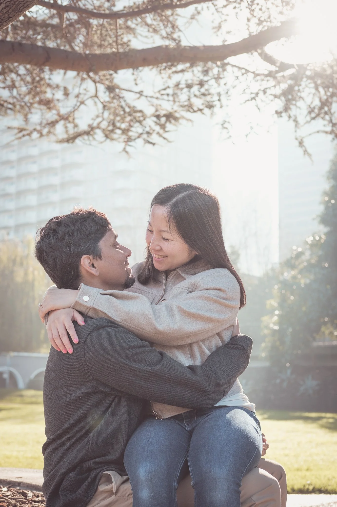 A couple embracing outdoors on a sunny day, smiling at each other, with a cityscape and trees in the background.