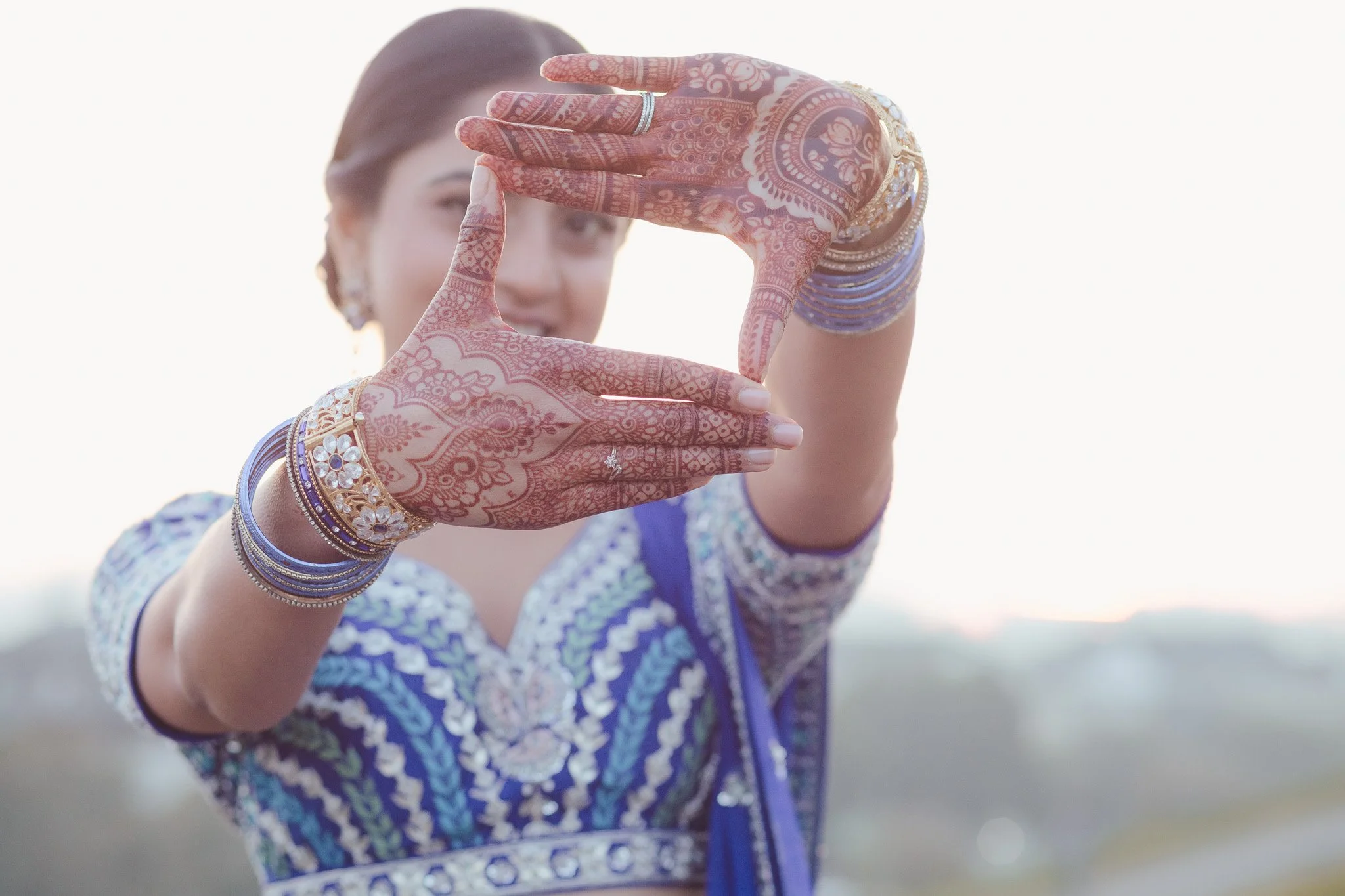 A woman dressed in traditional Indian attire holding her hands in front of her to frame her face, with intricate henna designs on her hands and gold jewelry, including bangles and rings, against a bright outdoor background.