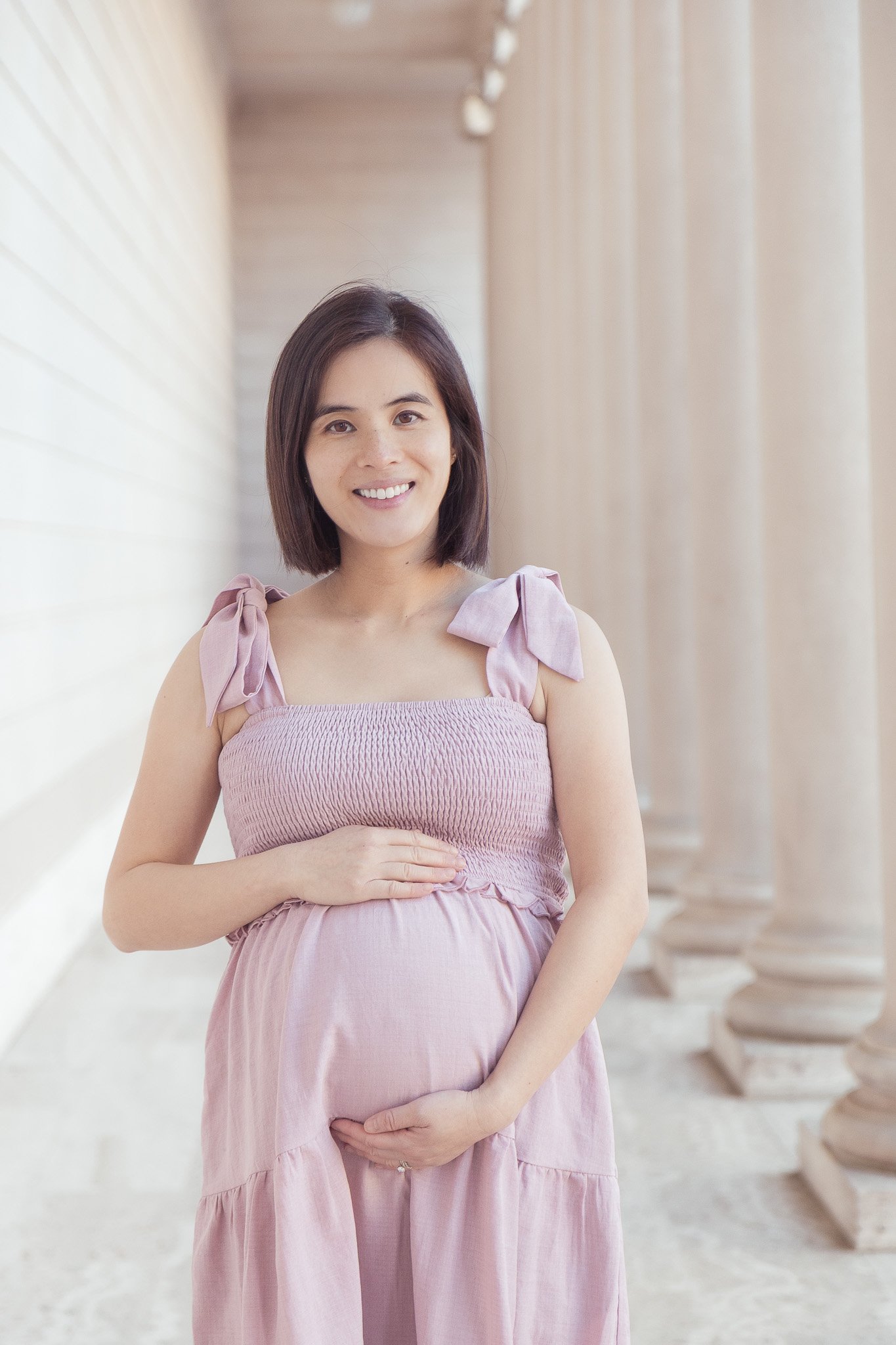 A pregnant woman in a pink dress standing outdoors, smiling, with one hand on her belly and the other resting on her stomach. She is next to large white columns and a light-colored wall.