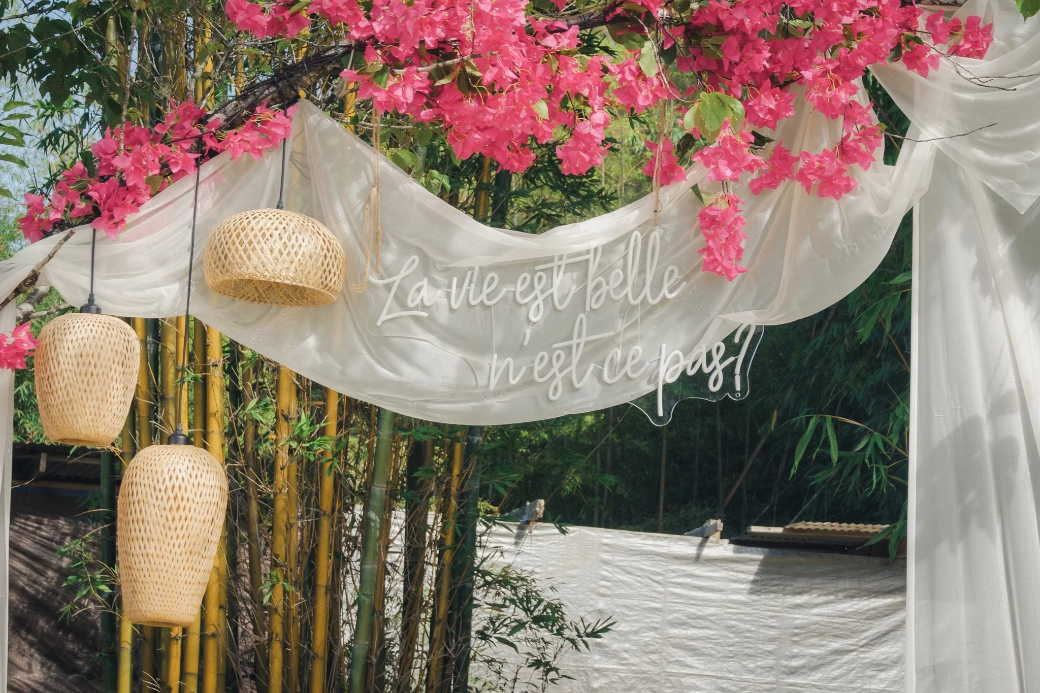 Outdoor decorated area with pink flowers, white fabric drapes, wicker lamps, and a white sign with French writing, set against a background of bamboo and greenery.