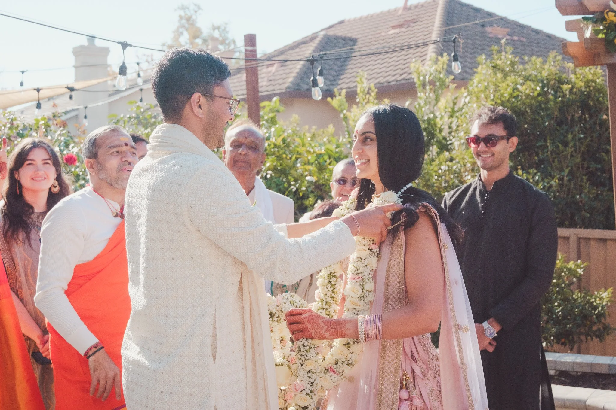 An outdoor Indian wedding ceremony with a bride and groom exchanging garlands, surrounded by friends and family.