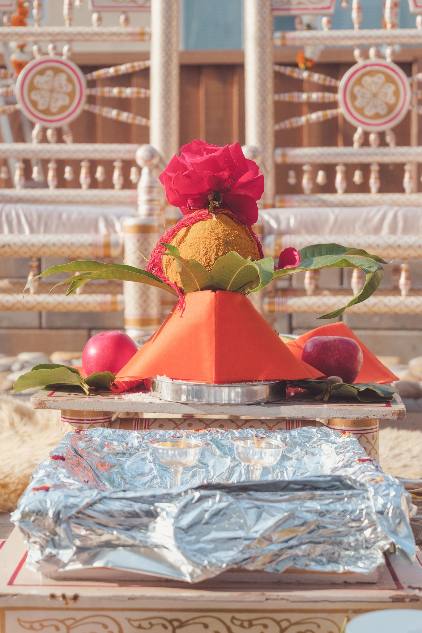 Hindu altar with a decorated banana fruit, red flowers, red apples, foliage, and candles, set against a backdrop of ornate chairs.