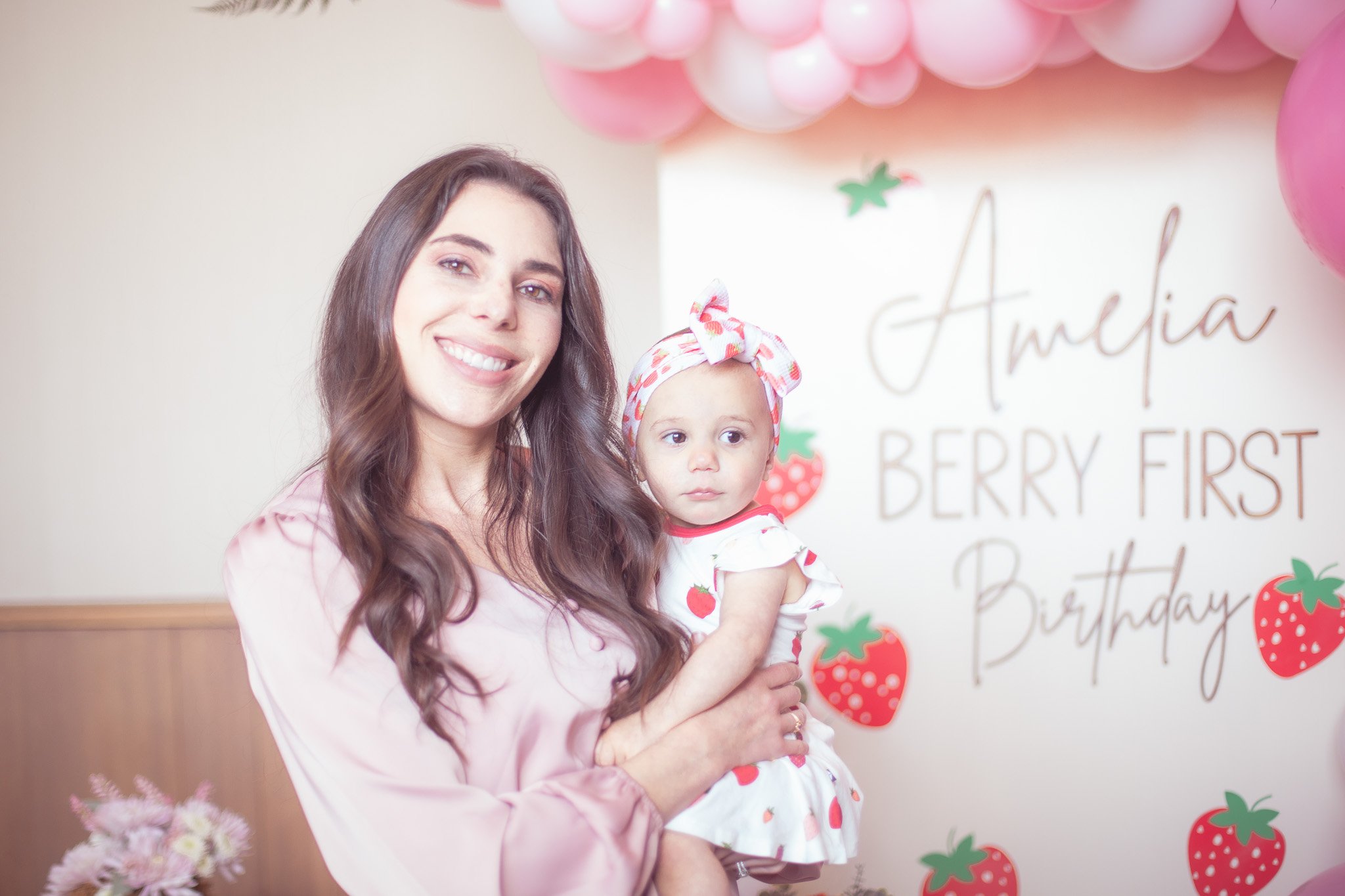 A woman smiling and holding a young girl at a birthday party with strawberry-themed decorations.