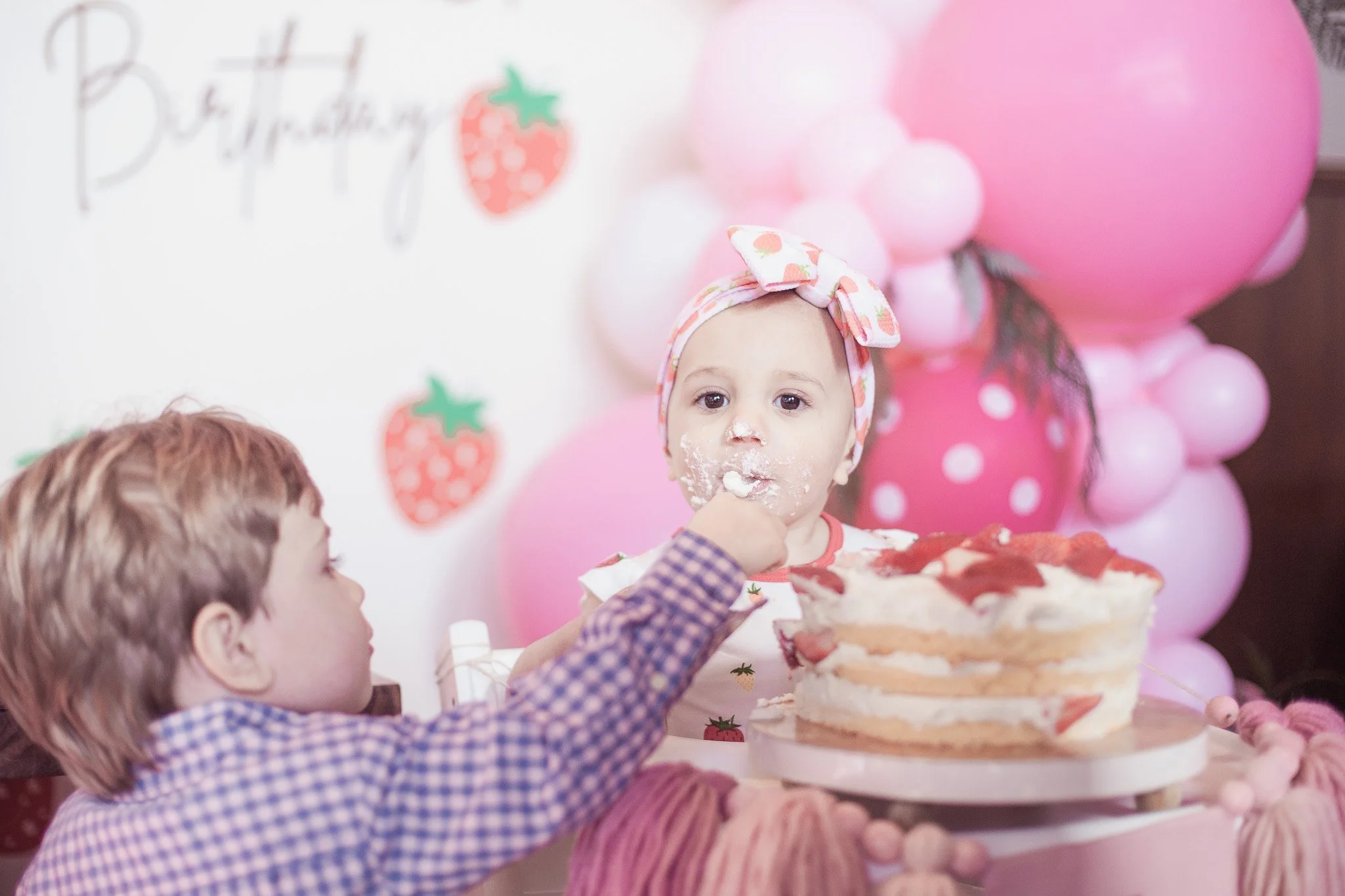 A young girl with a strawberry-patterned headband eating cake at a birthday party, with a boy helping her. The background features pink balloons and a strawberry-themed birthday decoration.
