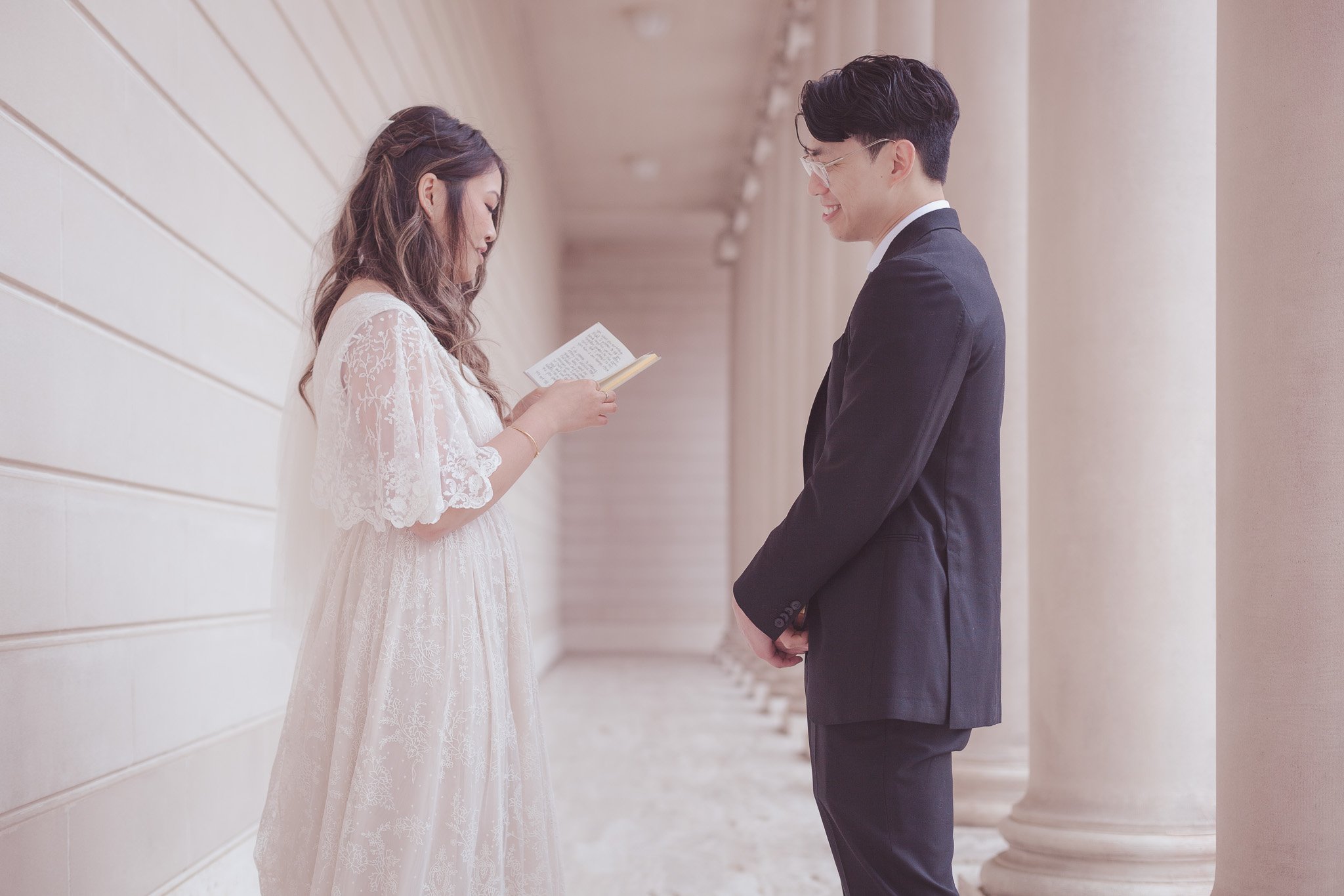 A couple standing in a column-lined hallway, with the woman reading from a small book as the man smiles and looks at her.