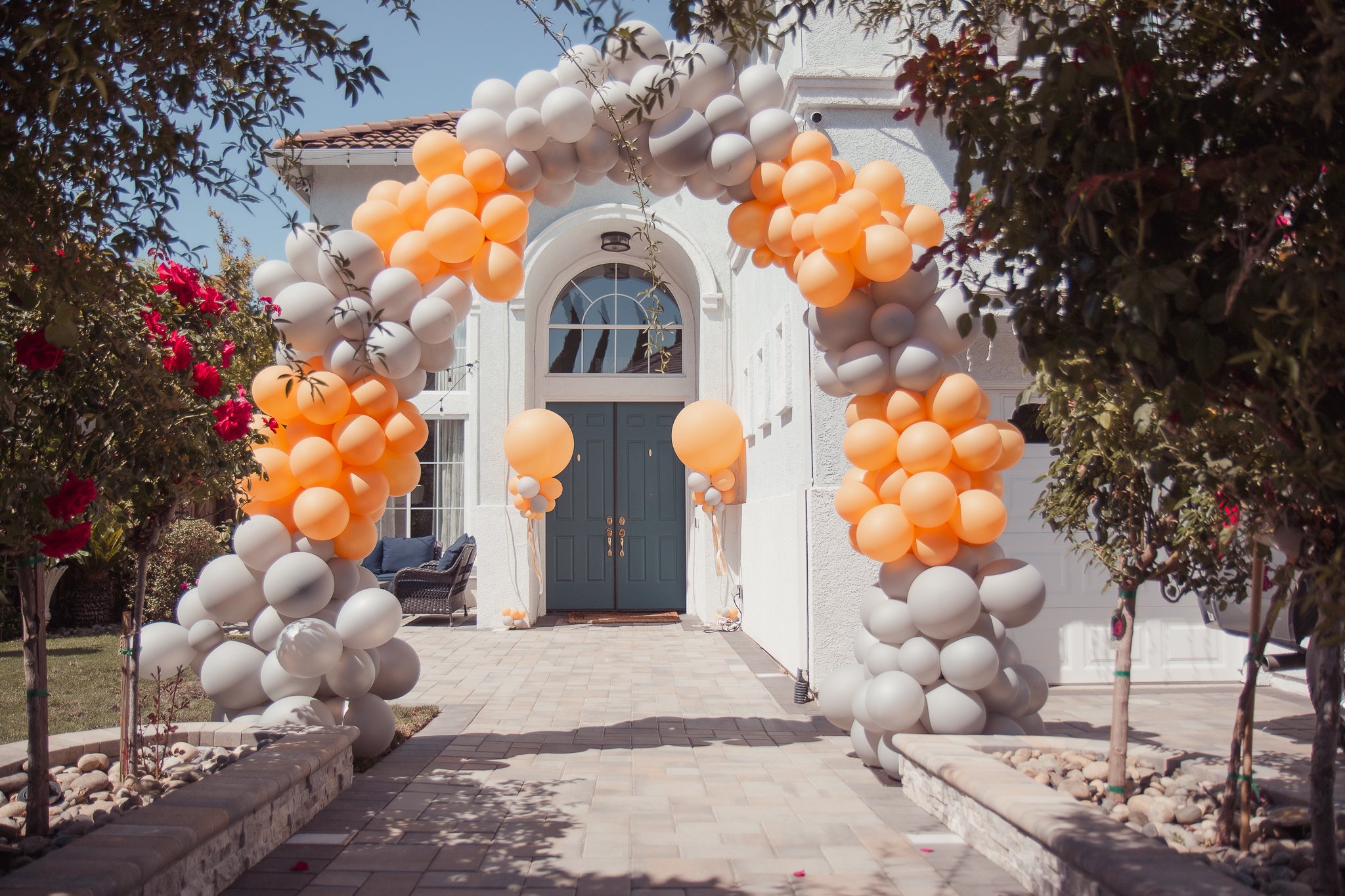Balloon arch with white and peach balloons decorating the entrance of a white house with a blue door.