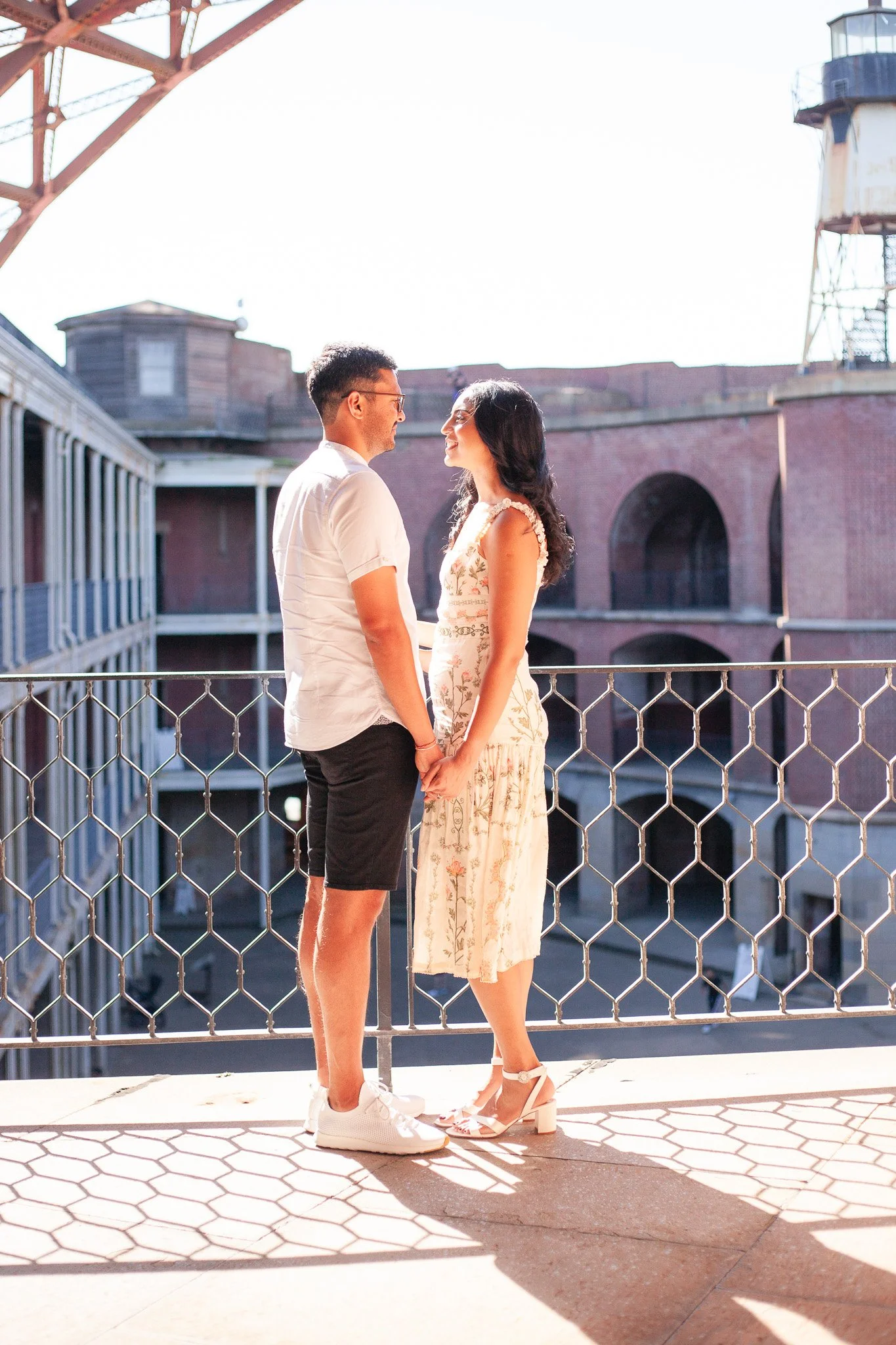 A couple holding hands and looking at each other on a balcony with metal railing, with brick buildings and a water tower in the background.