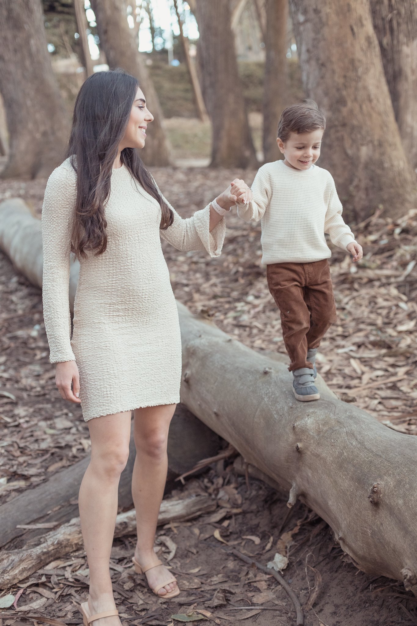 A woman and a young boy holding hands while walking on a fallen tree trunk in a wooded area.