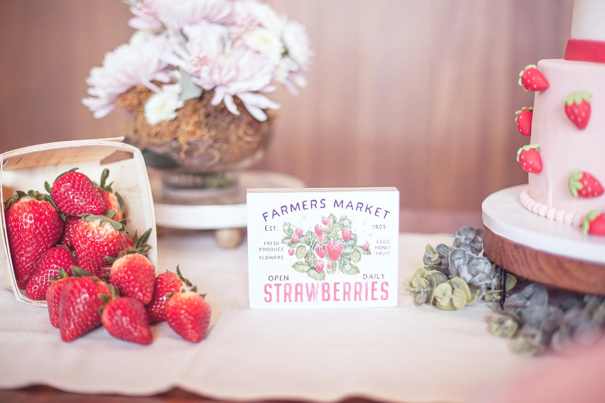 A table display featuring a basket of fresh strawberries, a vase with pink flowers, a sign for a farmers market selling strawberries, and a pink strawberry-themed cake with strawberries on its side.