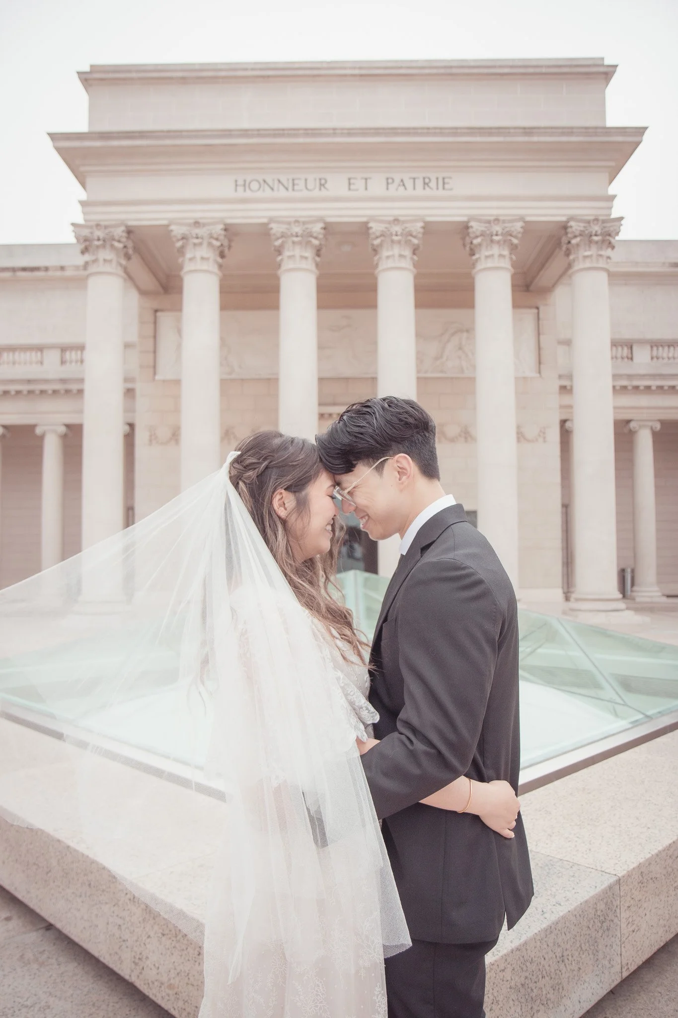 A bride and groom standing close with foreheads touching, smiling, in front of a historical building with columns and the inscription 'HONNEUR ET PATRIE'.