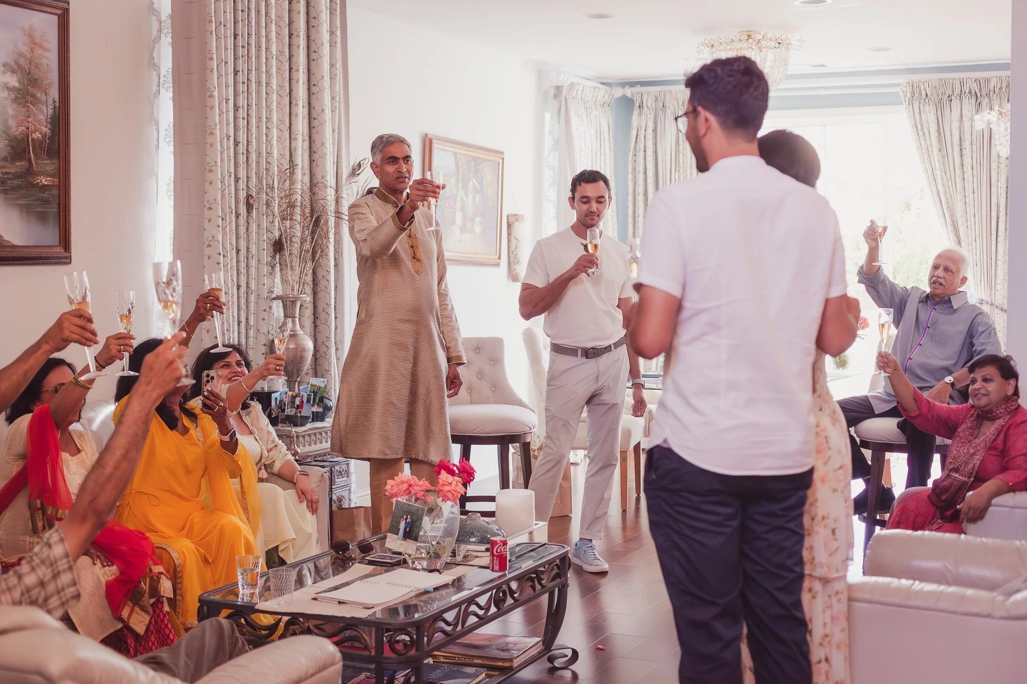 A group of people gathered in a well-lit living room celebrating with champagne, raising their glasses in a toast. The room has elegant curtains, a chandelier, and framed artwork.
