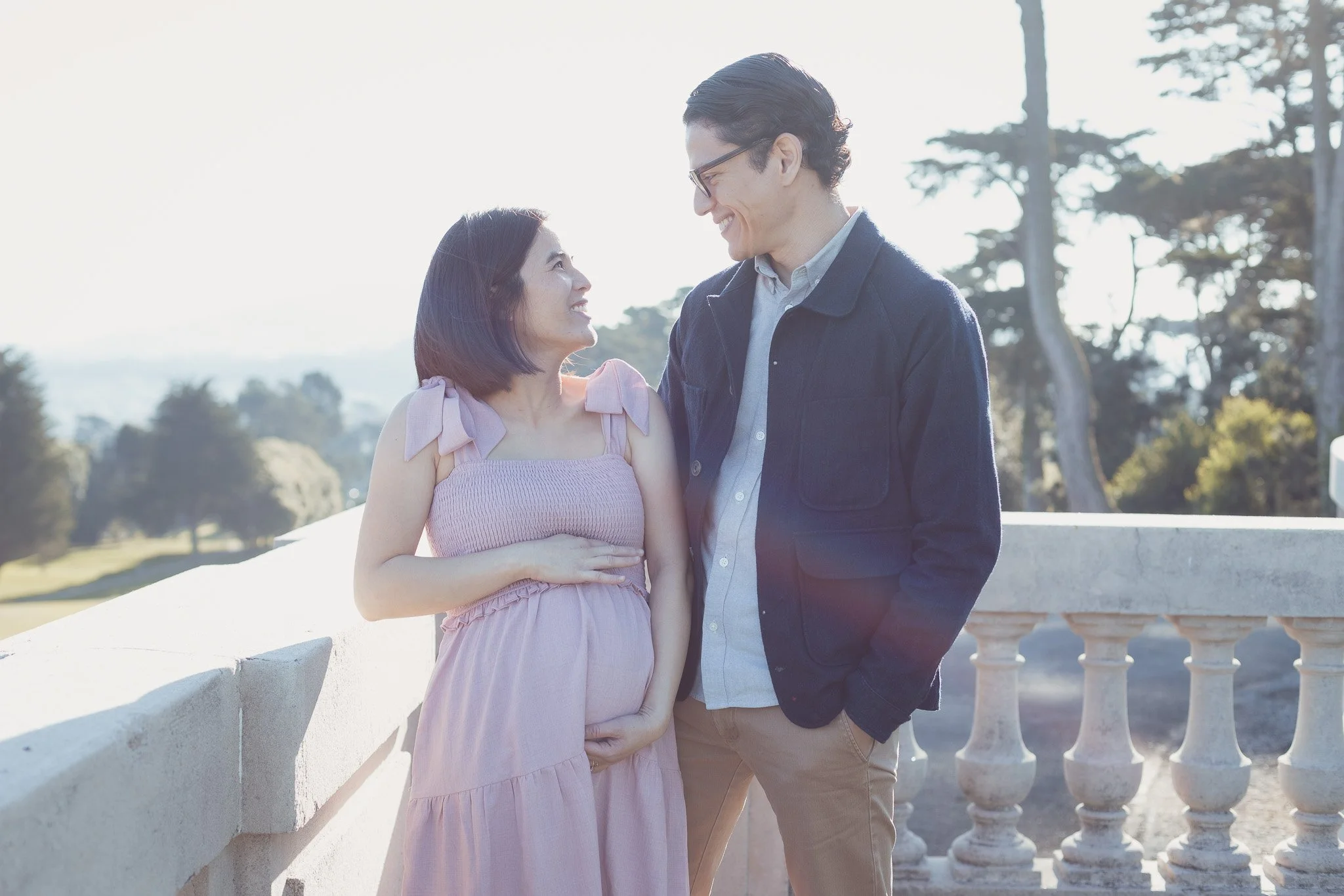 A pregnant woman and a man smiling at each other outdoors with trees in the background