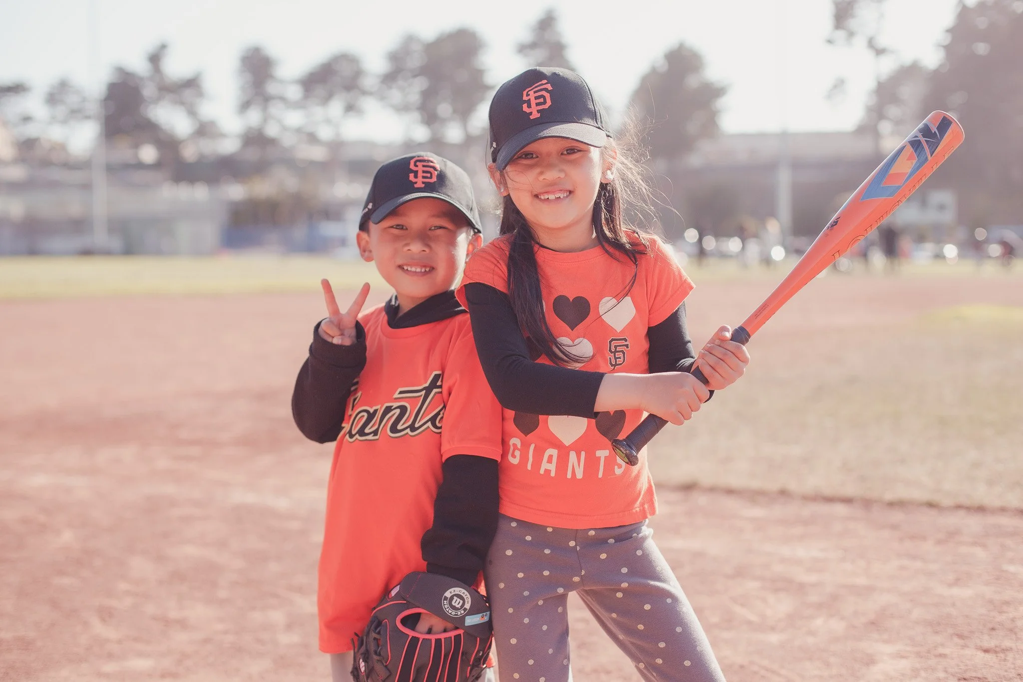 Two young children, a boy and a girl, wearing San Francisco Giants baseball caps and shirts, standing on a baseball field. The girl is holding a baseball bat, and the boy is making a peace sign with his fingers. They are smiling and posing together.
